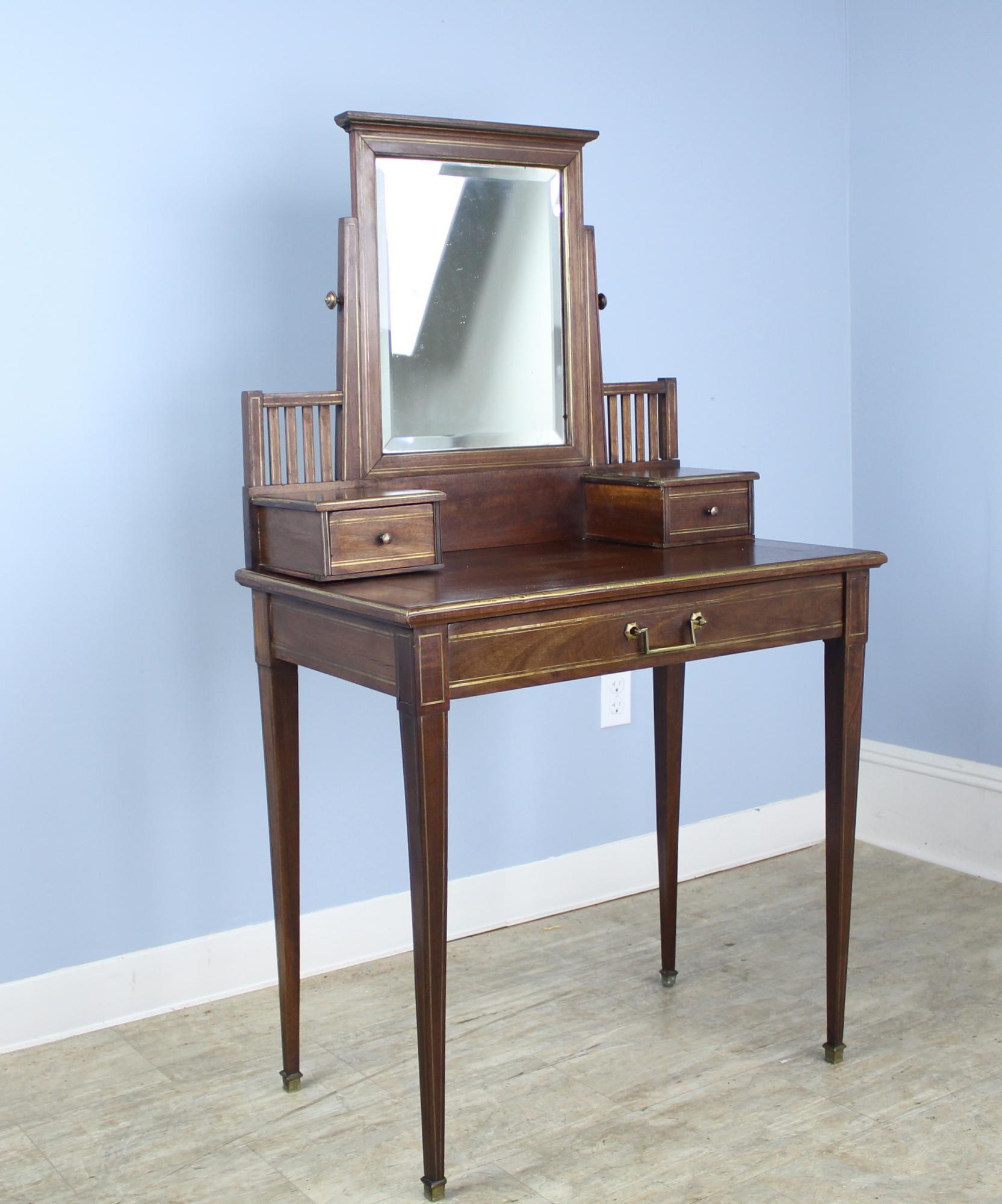 A dainty vanity or dressing table with brass details and three drawers, one wide single drawer in front and two smaller drawers for accessories. Original glass on the adjustable tilting mirror has some wear and a chip. Height measurement below is