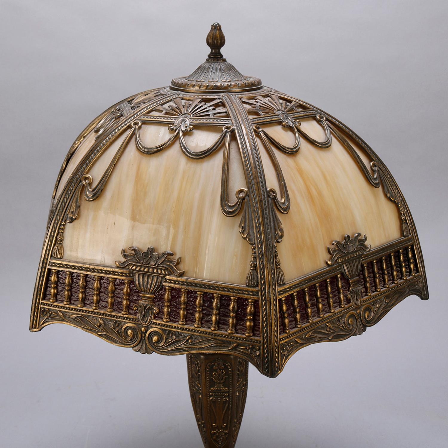 An antique Arts & Crafts two toned slag glass table lamp offers dome form shade with cast frame having stylized sunburst, foliate, urn, swag and tassel motif housing bent slag glass and surmounting faceted base, circa 1920

***DELIVERY NOTICE – Due