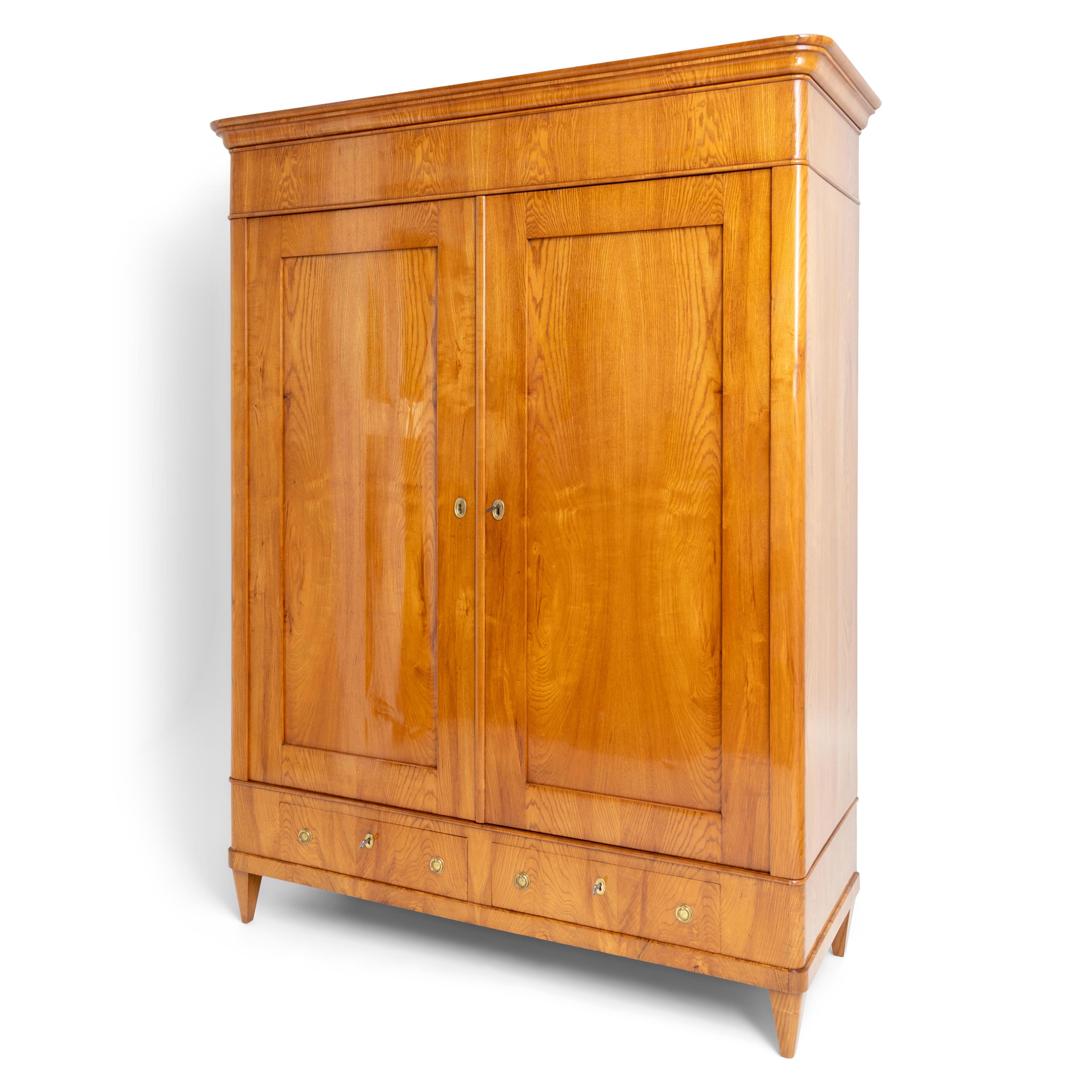 Large ash two-door wardrobe with profiled cornice, rounded corners and coffered doors and two drawers. The internal division consists of shelves and a clothes rail. Body width: 147 cm; body depth: 54 cm; interior depth: 49 cm. The cabinet has been