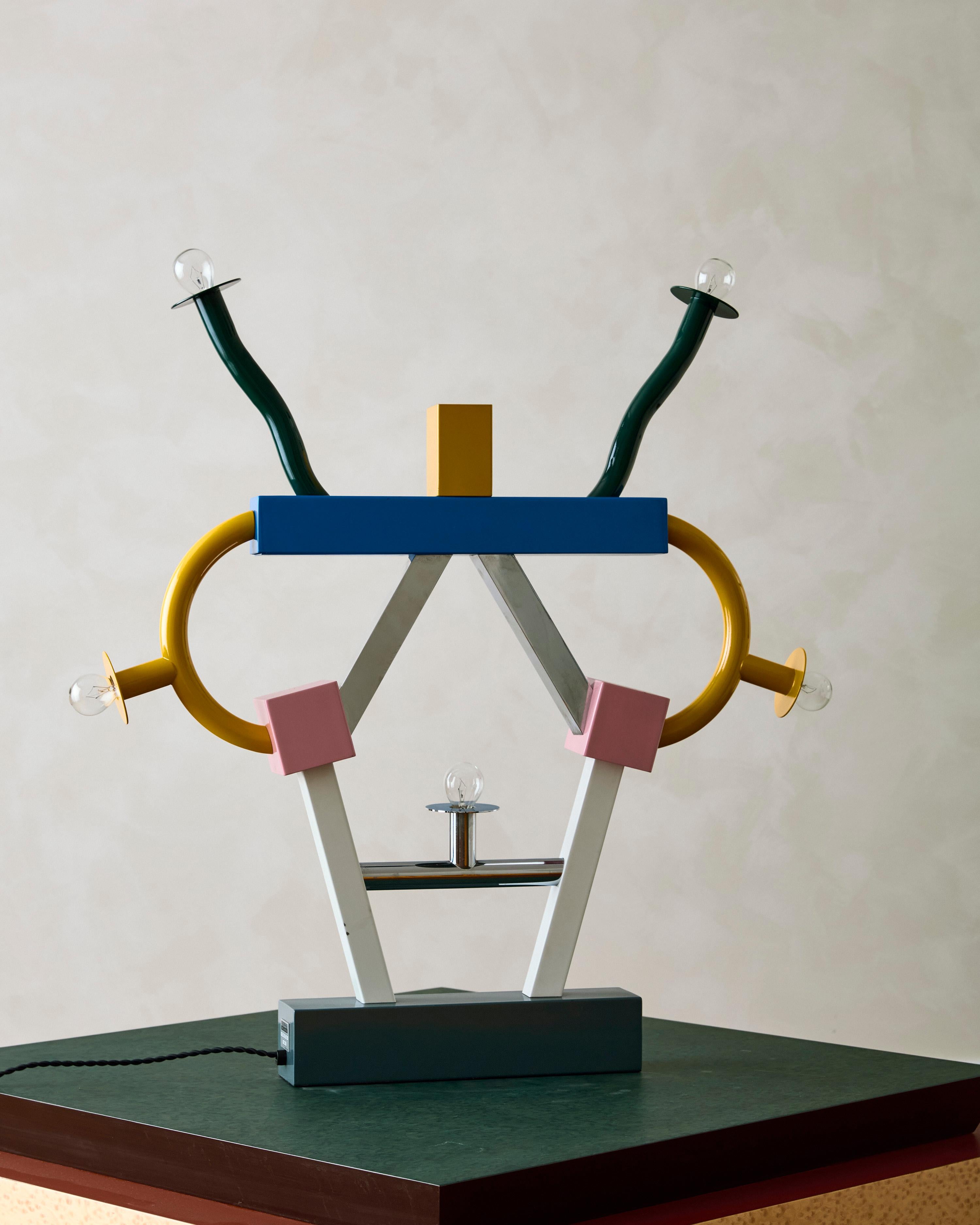 La lámpara de sobremesa Ashoka, diseñada por Ettore Sottsass para Memphis Milano, toma su nombre del emperador del Imperio Maurya. La lámpara está fabricada en materiales esmaltados y cromados, y presenta diversas formas geométricas.

Dimensiones: