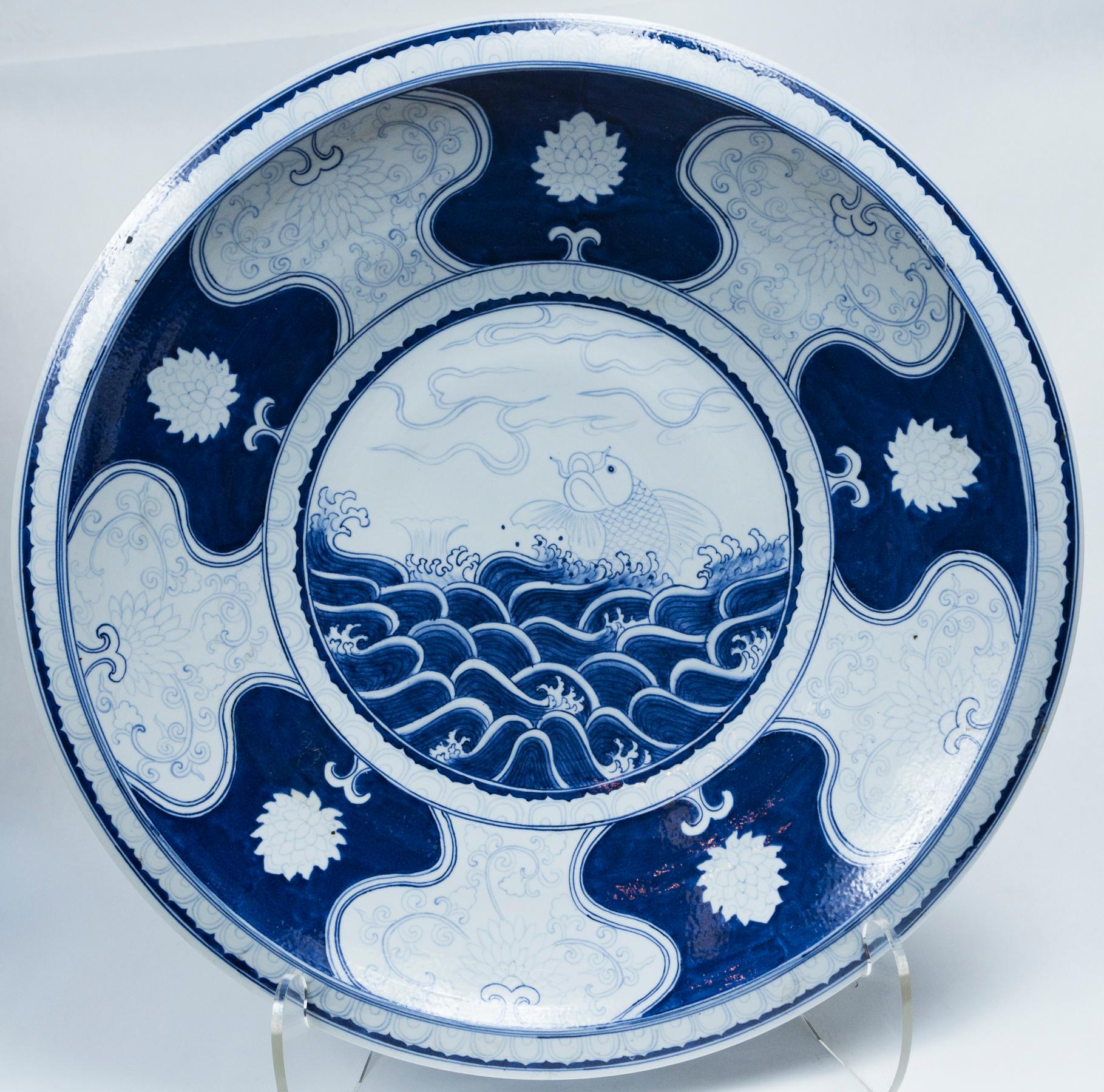 This charger or possibly a shallow bowl is centered with a leaping carp or koi amongst ocean waves. That is surrounded by 5 blue plaques with a chrysanthemum flower in white, the back rim with more intricate design in white, on a blue