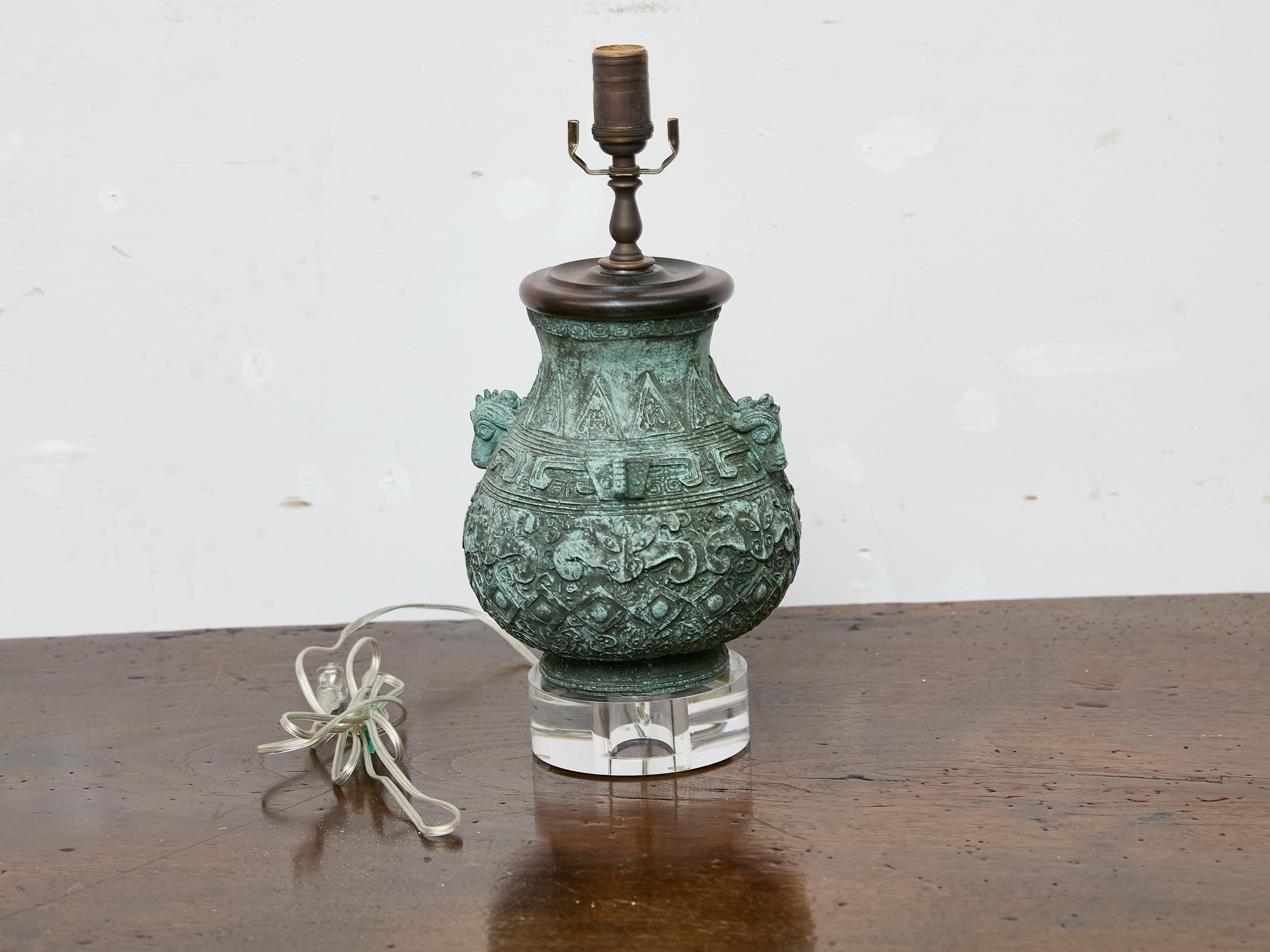 This distinctive table lamp is crafted from an early 20th-century Asian bronze vessel, mounted on a modern clear lucite base. The original vessel, dating to circa 1900, features an intricate relief design throughout, with stylized mythical