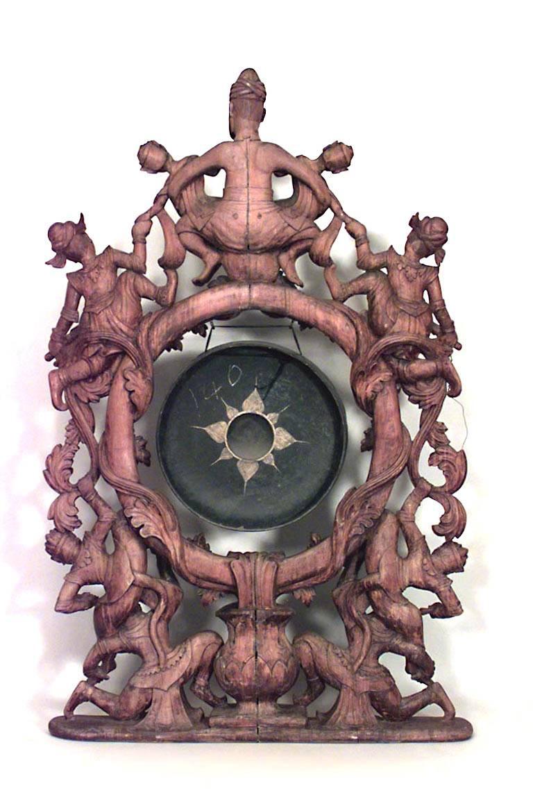 Asian Burmese style (19th century) large gong stand carved with figures of deities and demons with a centered hanging paint-decorated metal gong.
 