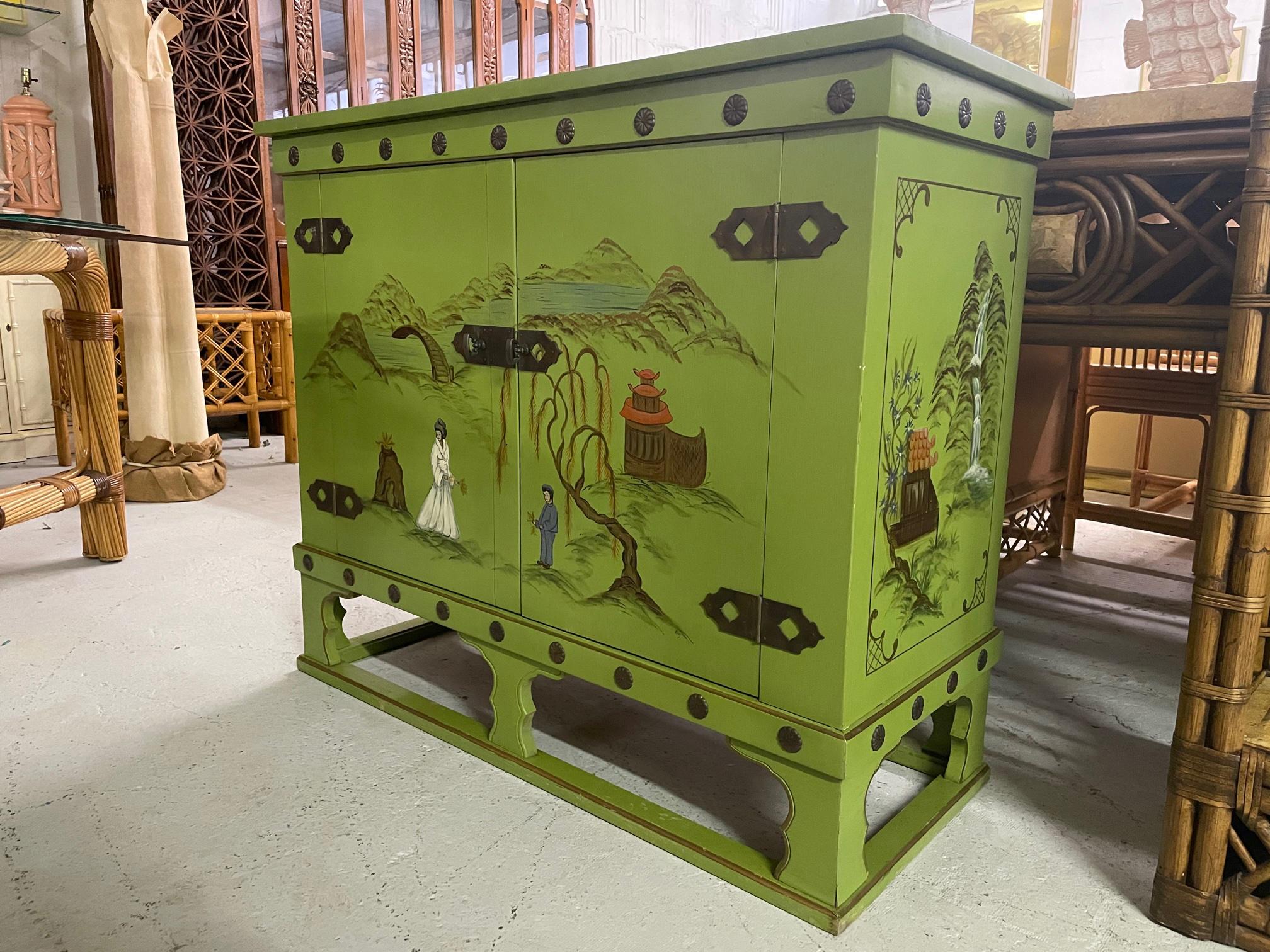 Asian Style bar cabinet features a beautiful green finish with hand painted Chinese scenes and brass hardware. Opens on top as well as double doors revealing storage for bottles and other barware. Good condition with minor imperfections consistent