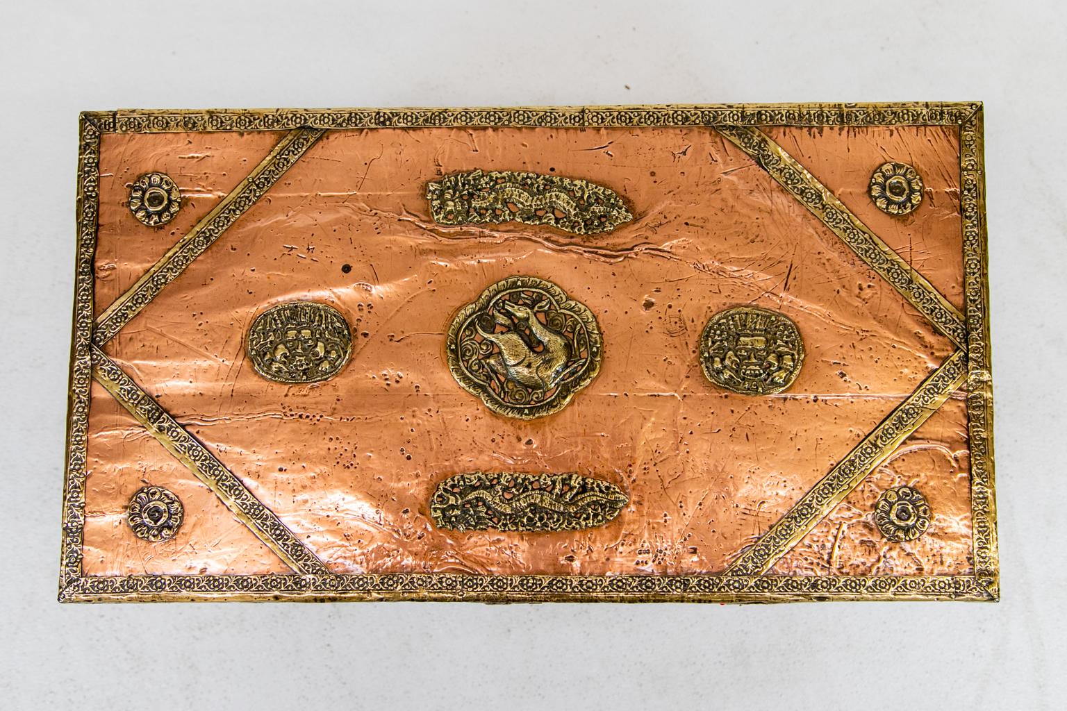 Le coffre asiatique revêtu de cuivre est cerclé d'une bande de laiton gaufré et de cartouches superposées. Des boutons rouges et bleus incrustés de rosettes se trouvent sur le devant et sur les côtés.
   