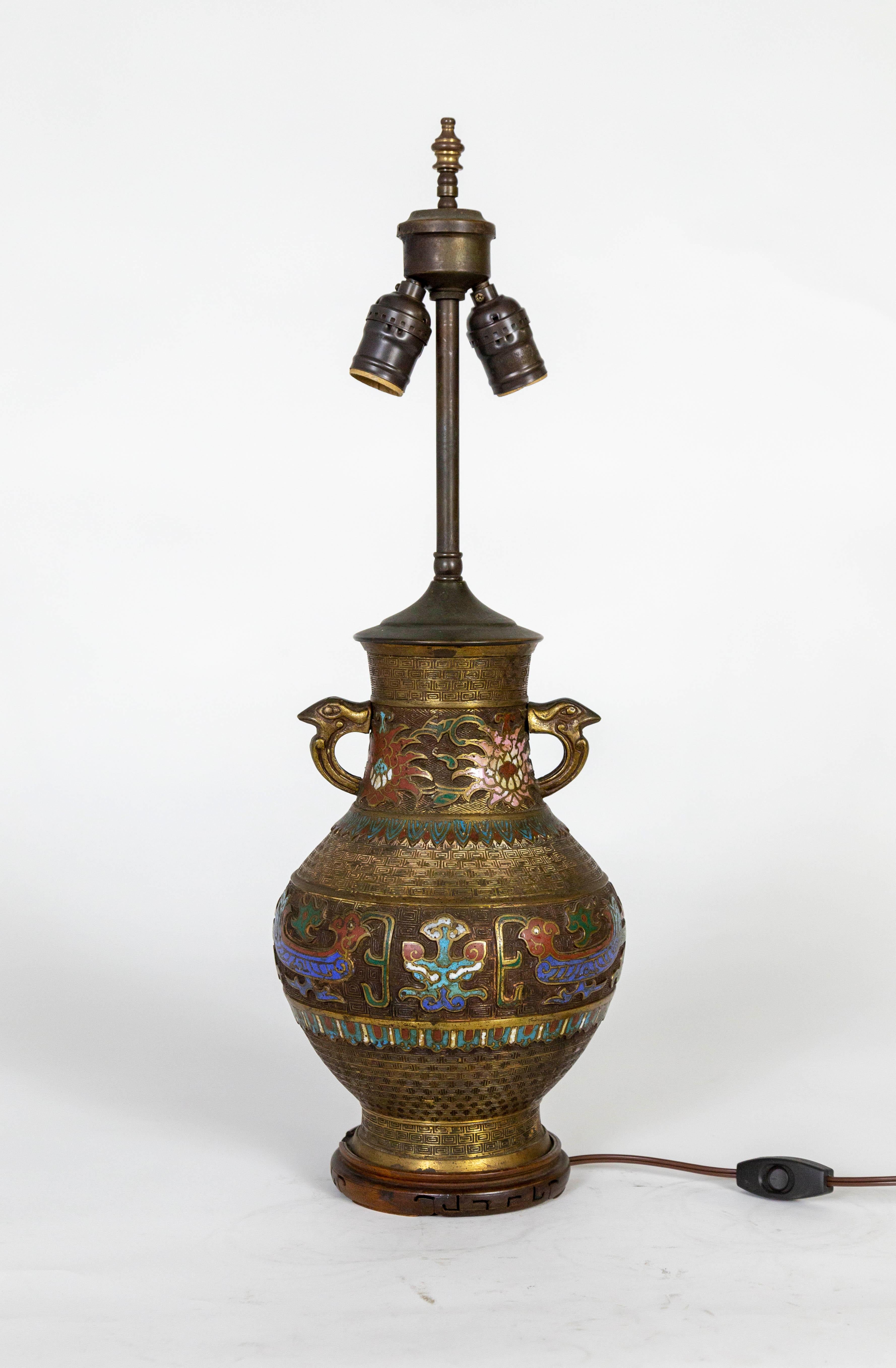 An early 20th century, patinated, cast bronze and champleve enamel vase that has been converted into a lamp; in an urn shape with an intricate greek key motif and handles cast with bird faces. Champleve is a technique that creates recesses in metal