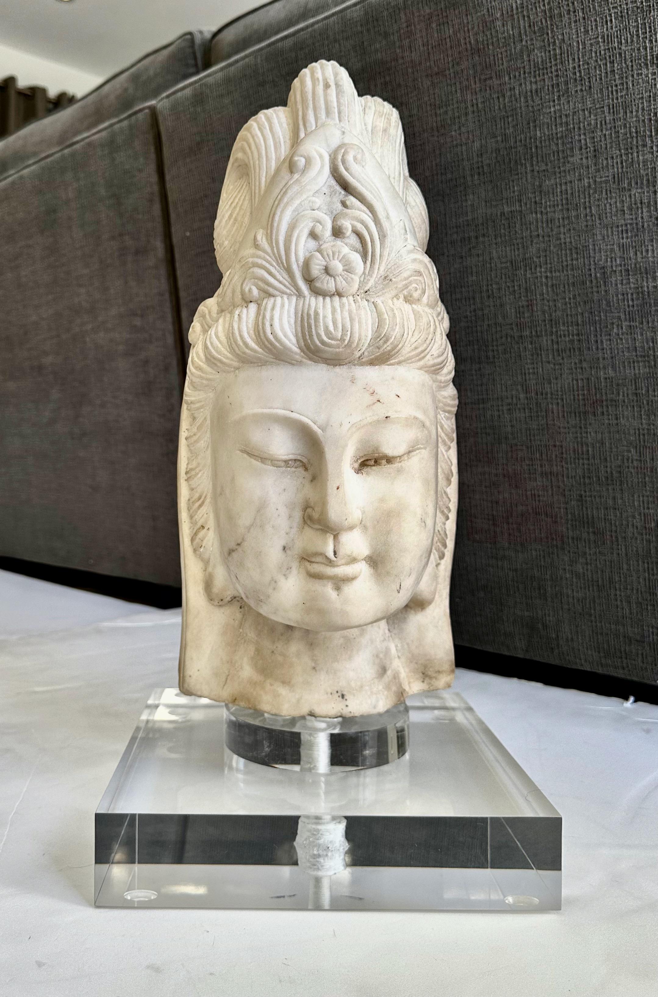 Beautifully executed hand carved marble buddha head fragment of Quan Yin. The youthful face is peaceful and serine framed by an intricately carved headdress. A striking art object with fine quality workmanship. Sits on custom acrylic base