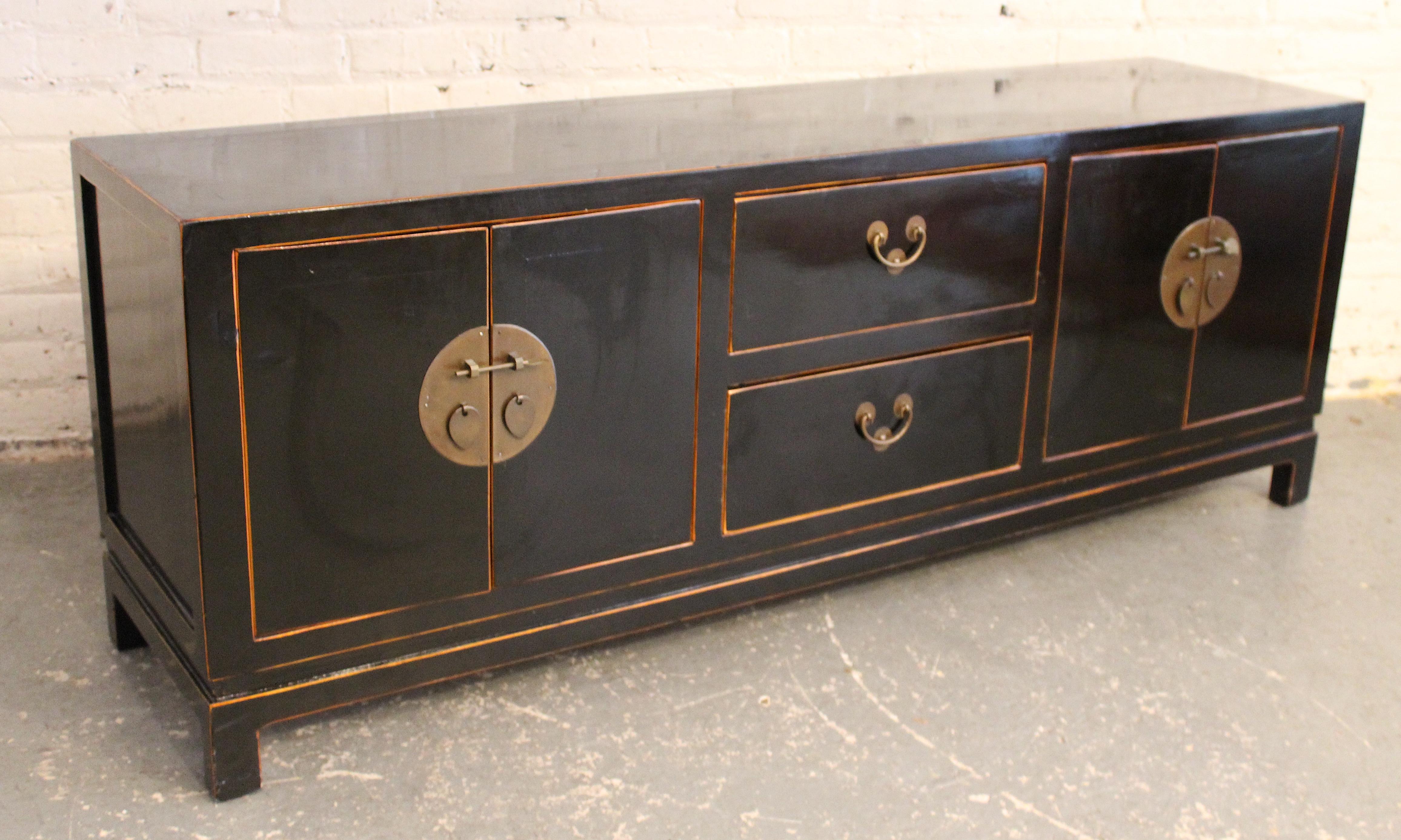 This low profile console cabinet, with Asian motif, is lacquered with a black piano style finish, and ornamented with brass hardware. Two flanking cabinets and middle drawer storage. 
Please confirm location NY or NJ