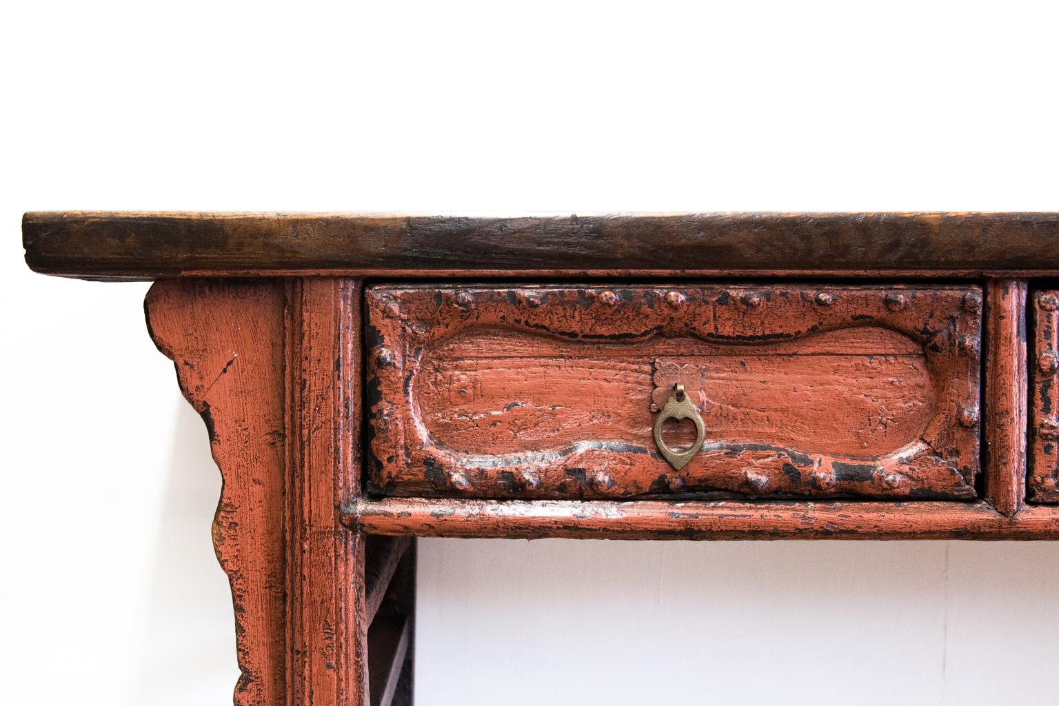 Asian painted console table with three drawers; the top has a waxed finish. There are carved moldings on the drawers and bordering applied to the edges of the drawers with engraved brass drop pulls. The legs are shaped, and there are double cross
