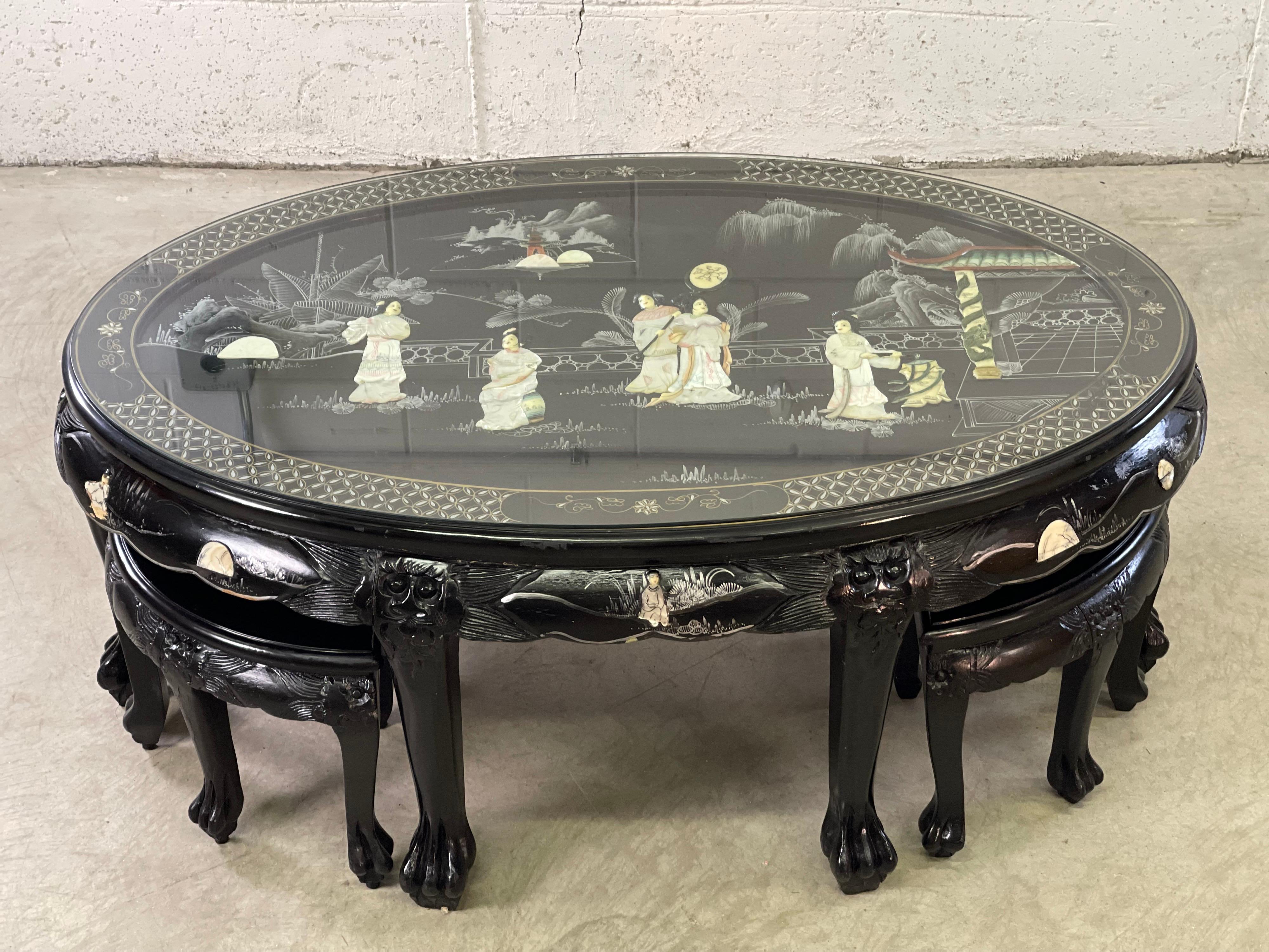 Vintage 1970s Asian style oval black lacquered coffee table with four stools. The table has multiple Asian scenes with bone and mother-of-pearl inlay. The table has carved claw feet. The table also has a glass top to protect the Asian scences. The