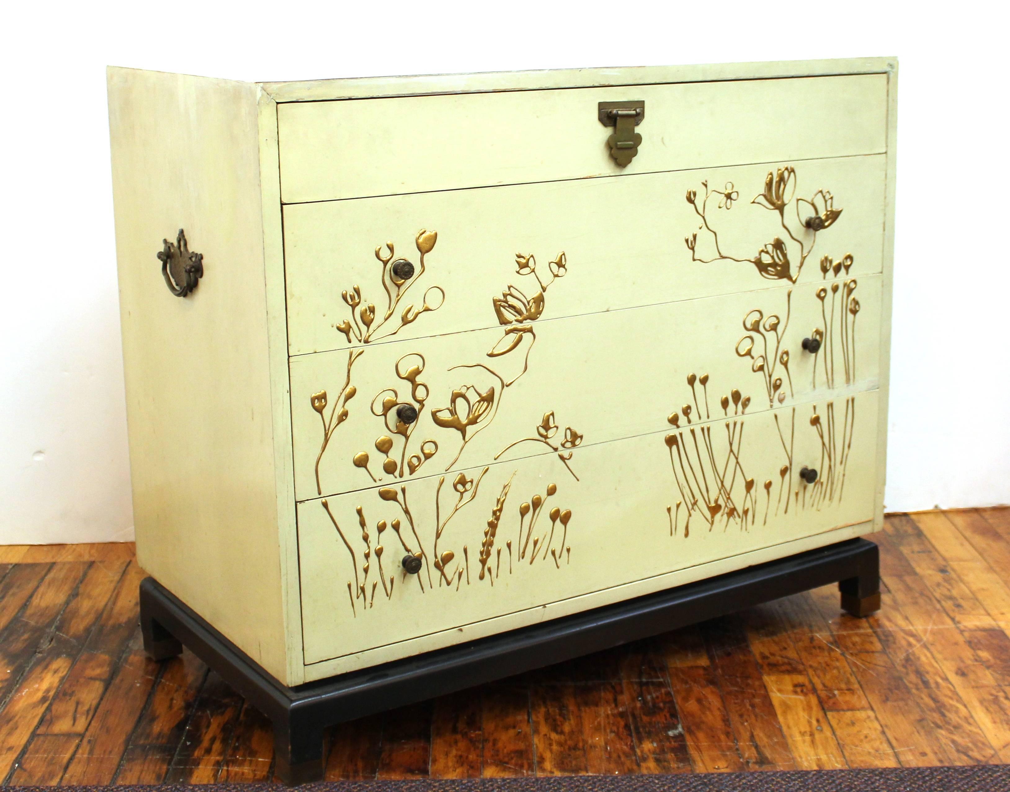 Asian style commode or chest with cream finish and decorative patinated brass hardware. Decorated with flowering plants hand painted in gold tone paint. The piece stands on a black base on block feet. Stamped on the back with description and serial
