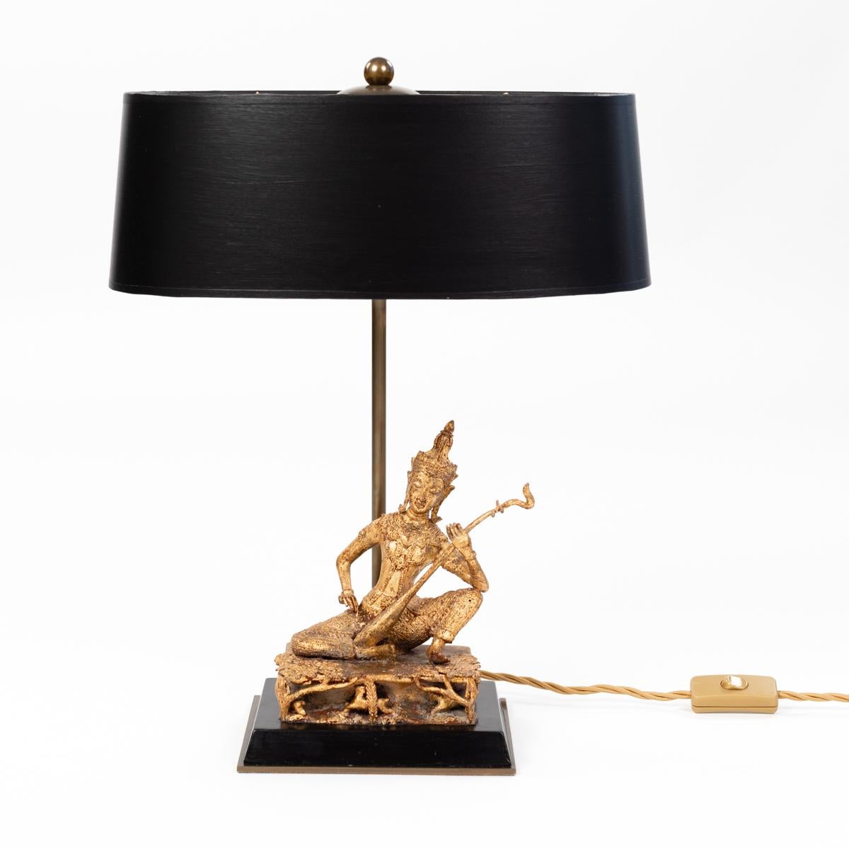 
Small figural table lamp of a seated temple musician in gilt bronze on a black wooden base. 
The noble object dates from the beginning of the 20th century.
Thai temple guardian sits on an ornate pedestal and plays a side instrument.
The figure is