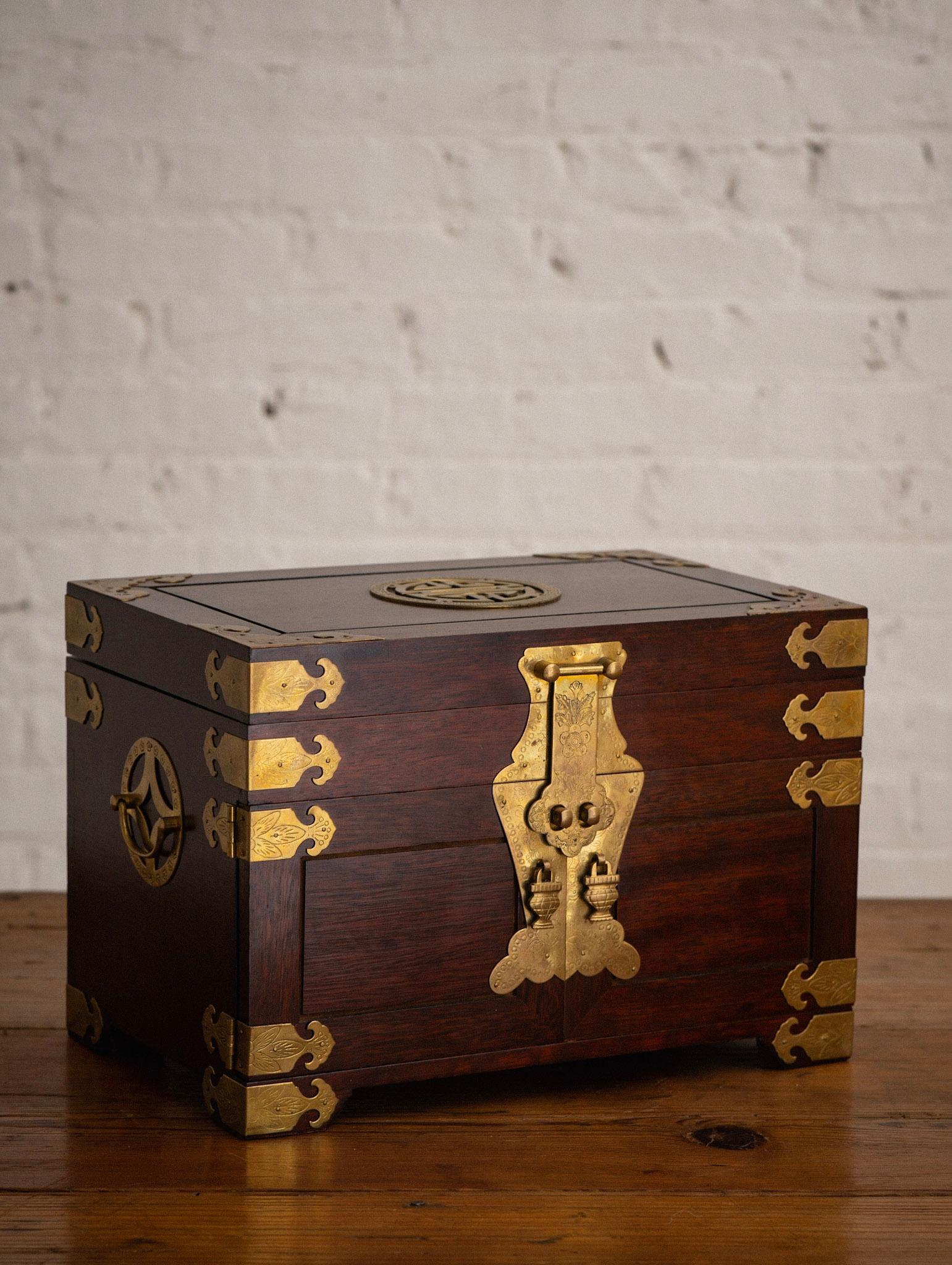 An asian wood and brass jewelry box. Dark red mahogany colored wood with with brass drawer pulls and decorative accents. Two doors open to reveal three inner drawers. Upper lidded compartment. Red floral interior. 