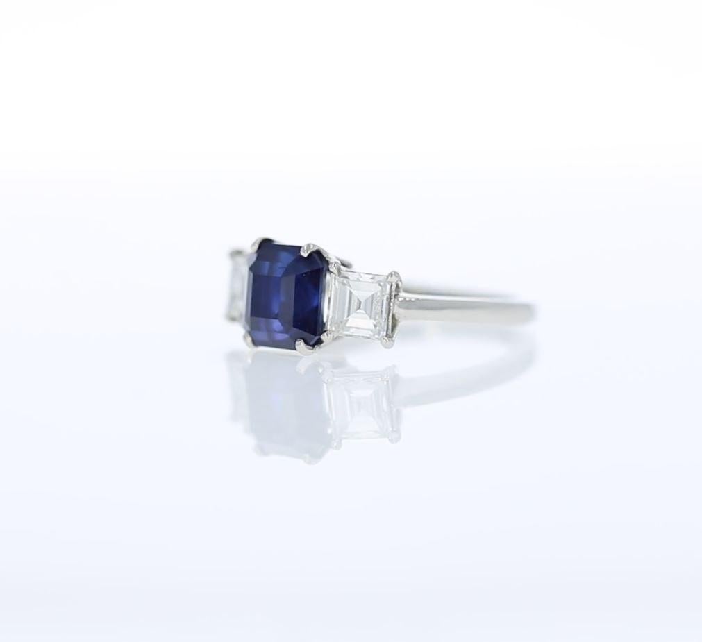 The rich royal blue of this Ceylon sapphire flashes its facets in a stunning Asscher cut, weighing a generous 3.31 carats. It is accompanied by two bright white square emerald-cut diamonds and set in gleaming platinum. The pair of diamonds together