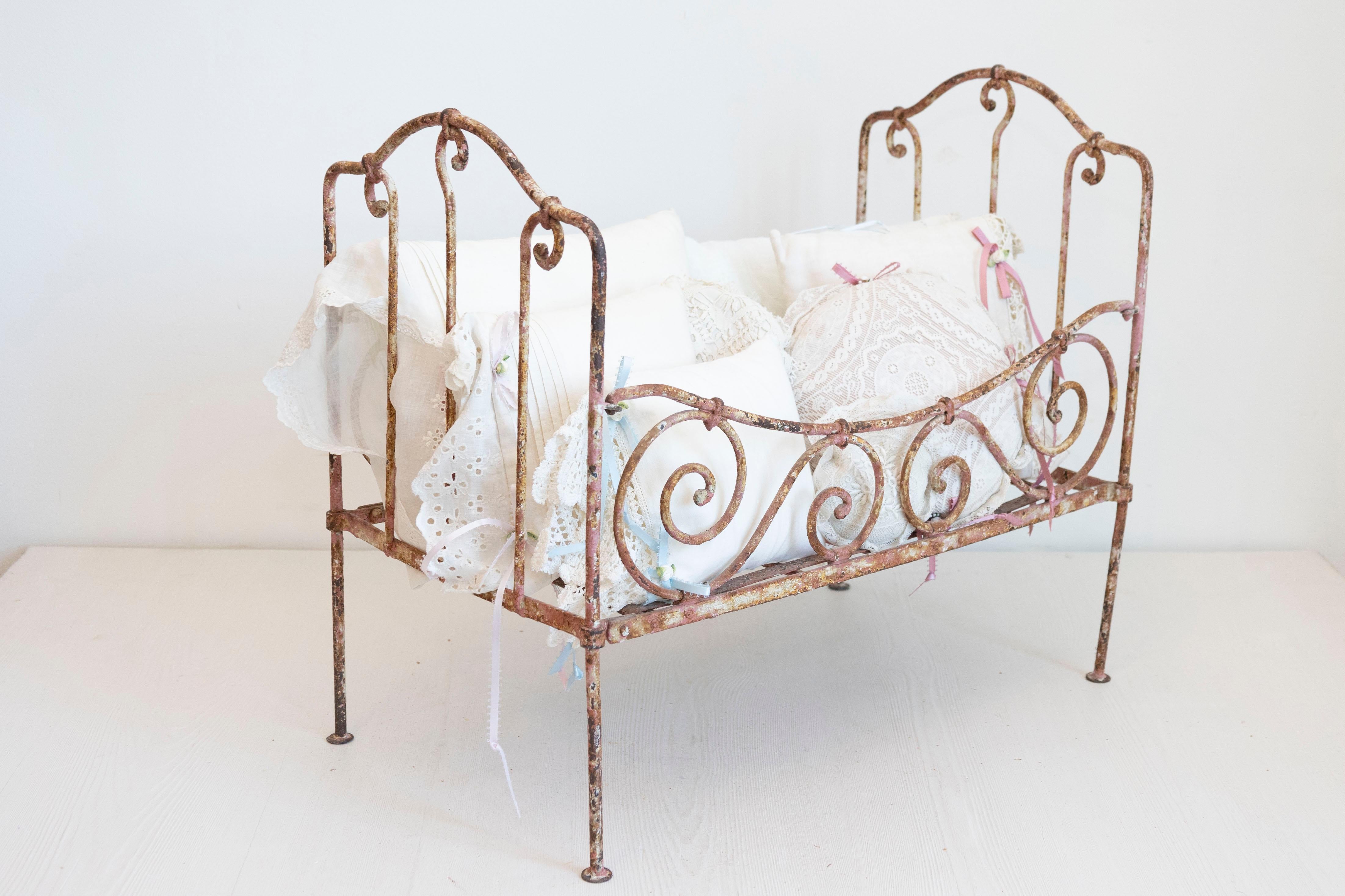 Surtido de almohadas vintage de encaje francés en cuna metálica del siglo XIX. Creada en Francia durante el siglo XIX, esta cuna petite cautiva nuestros corazones con su delicada disposición de cojines de encaje vintage atados con cintas azules y