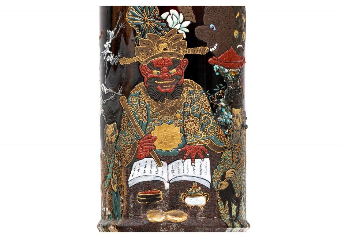 Cylindrical jars glazed with striated colors in the top background. With overall dense raised colorful decoration of a mythological scene with a variety of figures. A prominent figure with monstrous mask and headdress is seated reading from a book,
