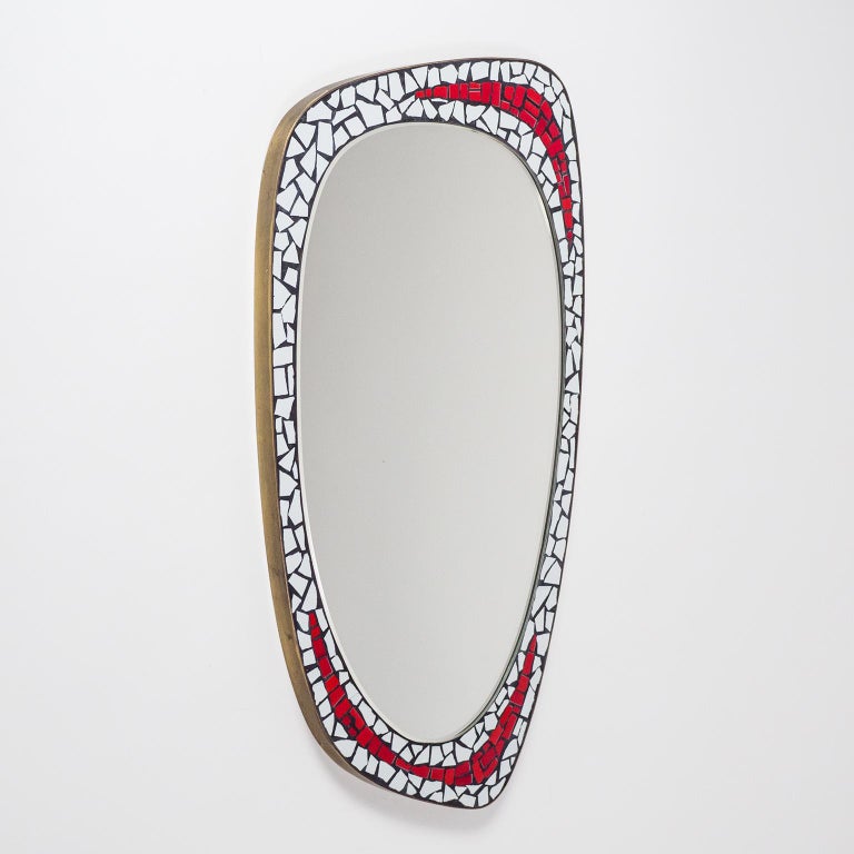 Charming midcentury mosaic mirror from the 1950s. An oval mirror is framed by an asymmetric ceramic mosaic in white and red with a solid brass rim. Fine original condition with minor wear to the mirror which measures 15inches/38cm tall and