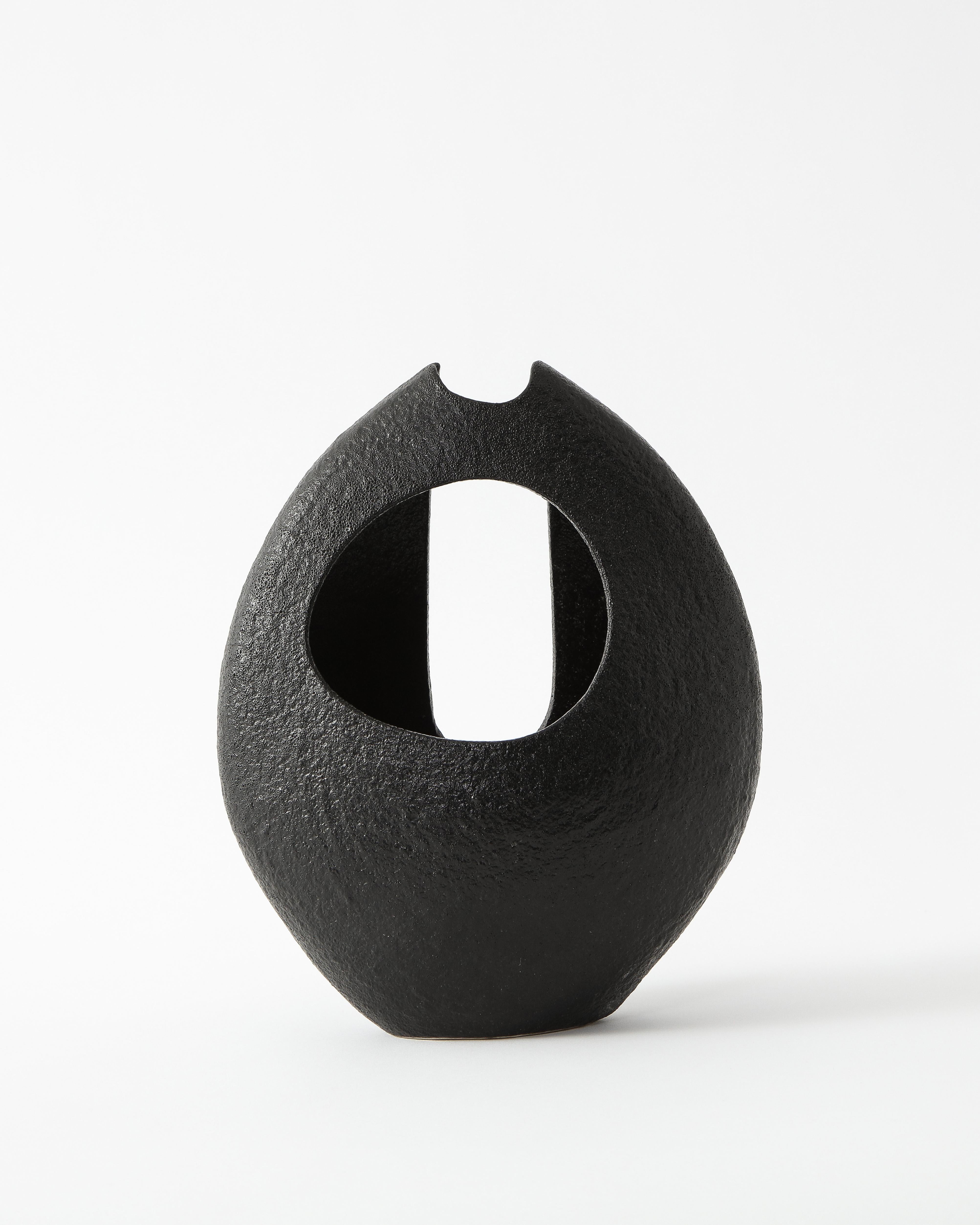 Asymmetrical black Japanese almond-shaped ceramic with a dense, grainy texture. One side features a circular hole while the other has an oblong opening that stretches up to the very top.