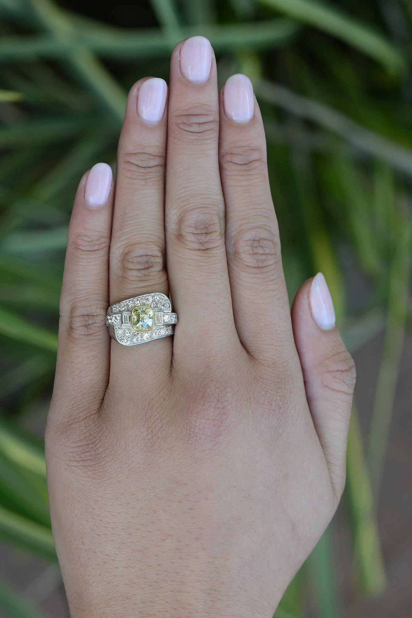 Nothing compares to an Art Deco engagement ring. Asymmetrical, curvaceous lines are shown throughout studded with shimmering old cut diamonds. At the center is a 1.42 carat old mine cushion diamond with a captivating, yellowish green with chunky,