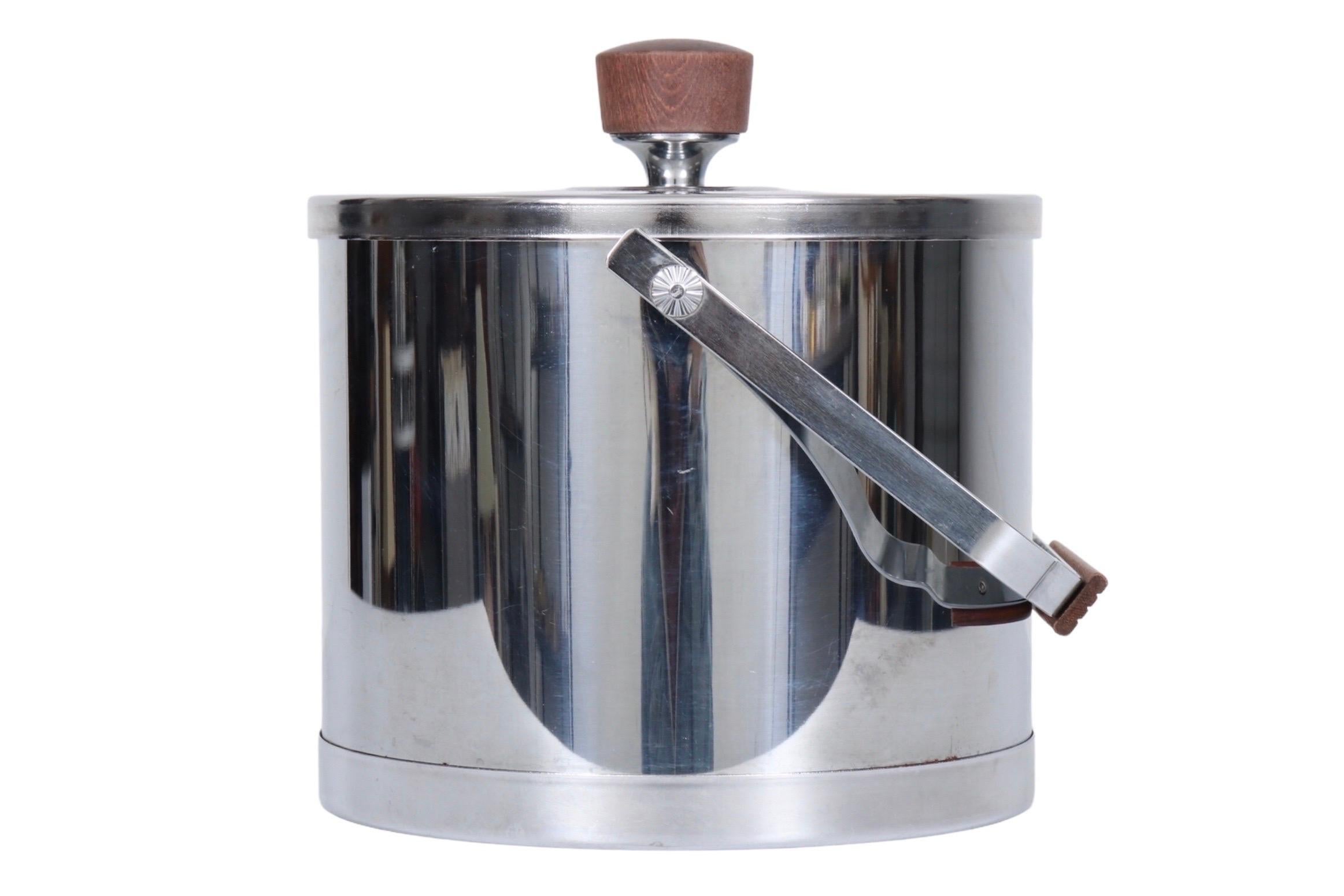 A 1970s mid century modern ice bucket by Atapco. Made of stainless steel with a squared steel handle topped with a teak bar handhold with reeded details. The lid lifts off with a teak knob handle and inside is a plastic liner. Marked 