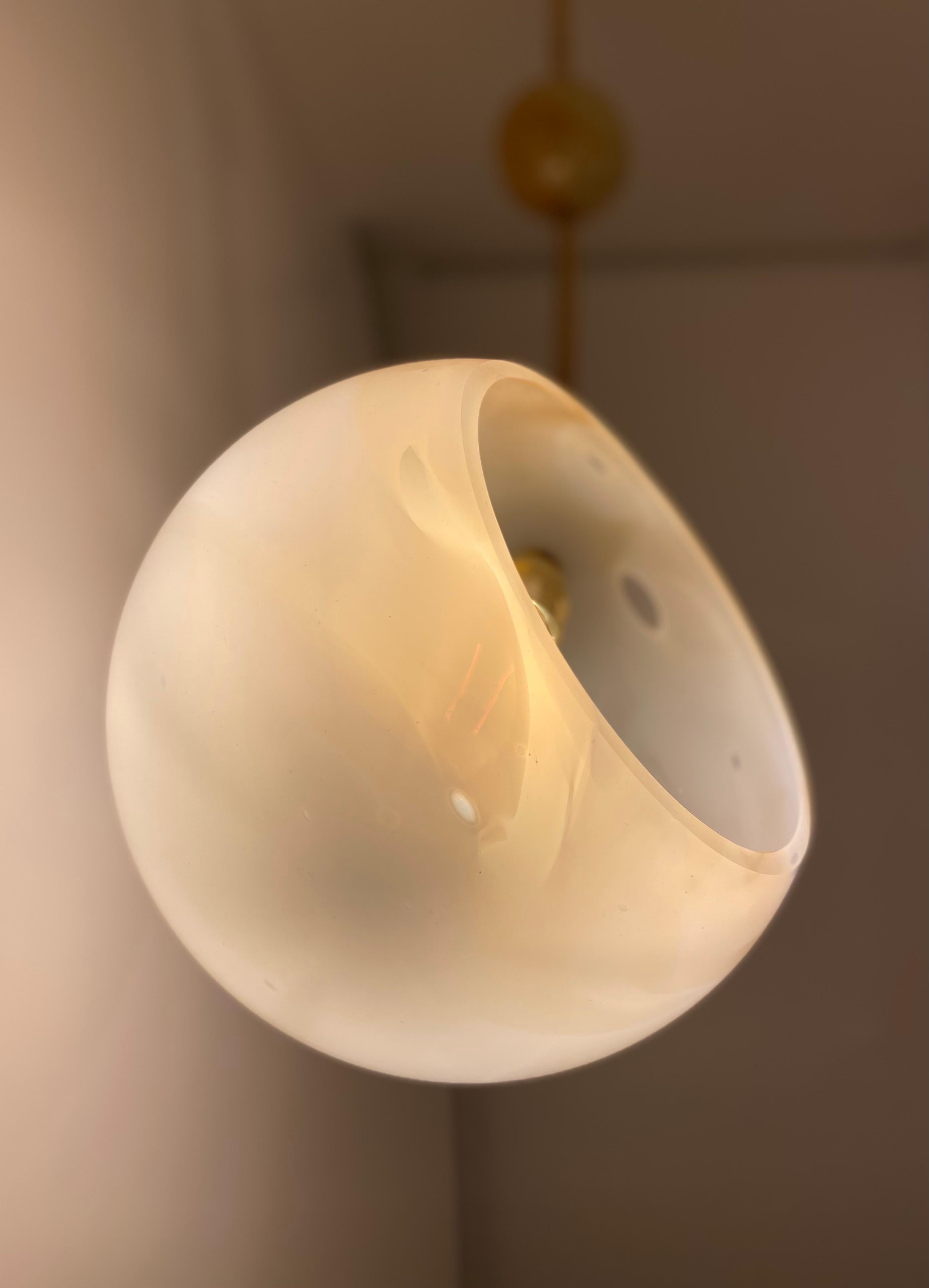 Die Solar-Pendelleuchte von Atelier George ist ein Einzelstück aus mundgeblasenem Glas mit einem Messingstiel. Sie besteht aus einer weißen Glaskugel mit bernsteinfarbenen Adern und drei Glasplaneten in blauen, grauen und ockerfarbenen Tönen, die