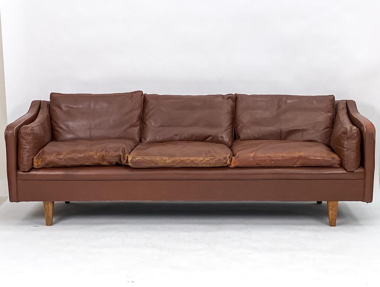 A fabulous Danish mid-century three-seater sofa in rich brown handsomely patinated leather. Attributed to Illum Wikkelso, this Scandinavian modern sofa has an attractive refined design with gently sloping armrests and triangular legs in teak wood,