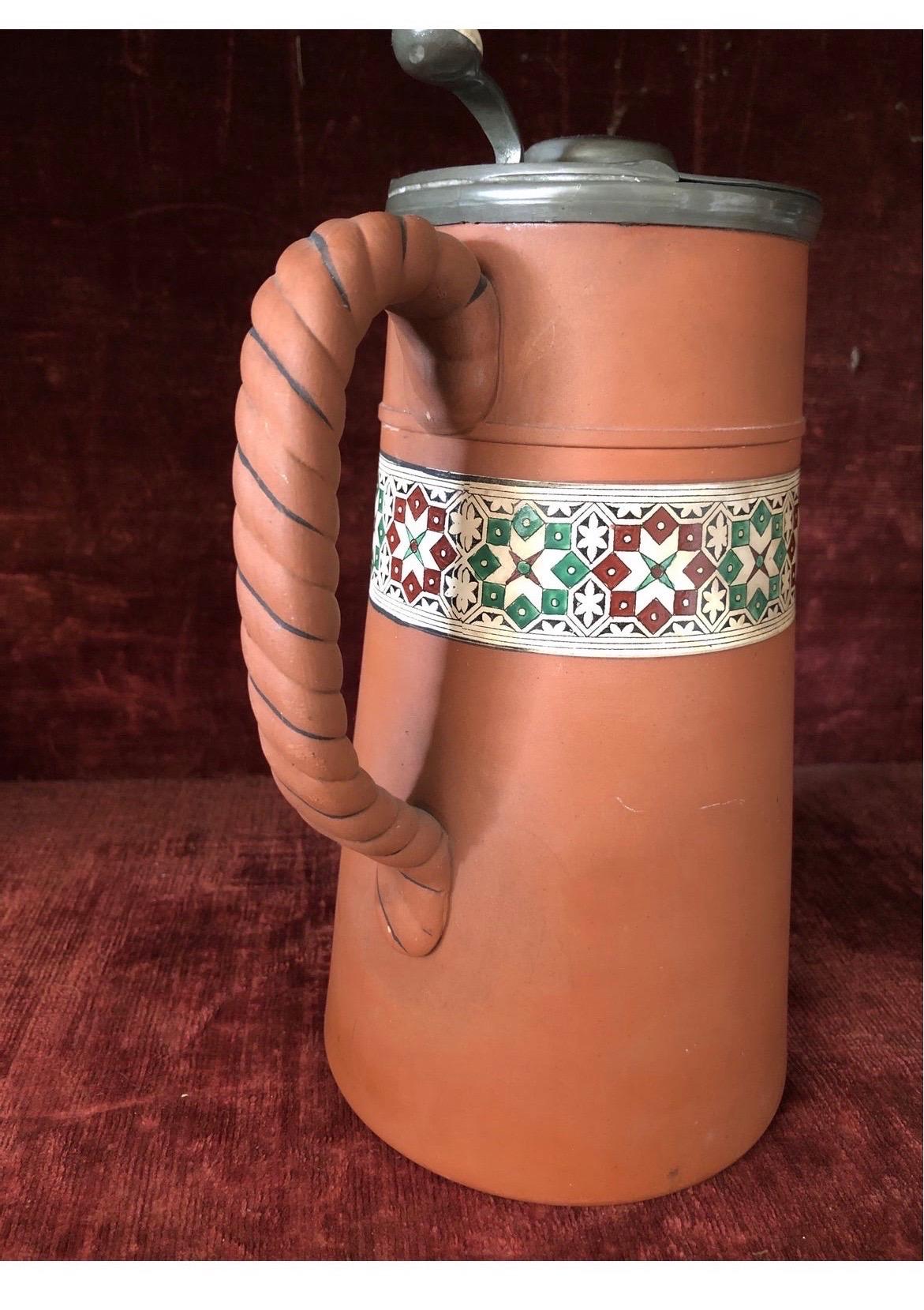 A good quality antique terracotta pitcher with hinged lid, twist braided handle and enamel decoration to center. 
No maker mark present, however, likely Wedgwood.