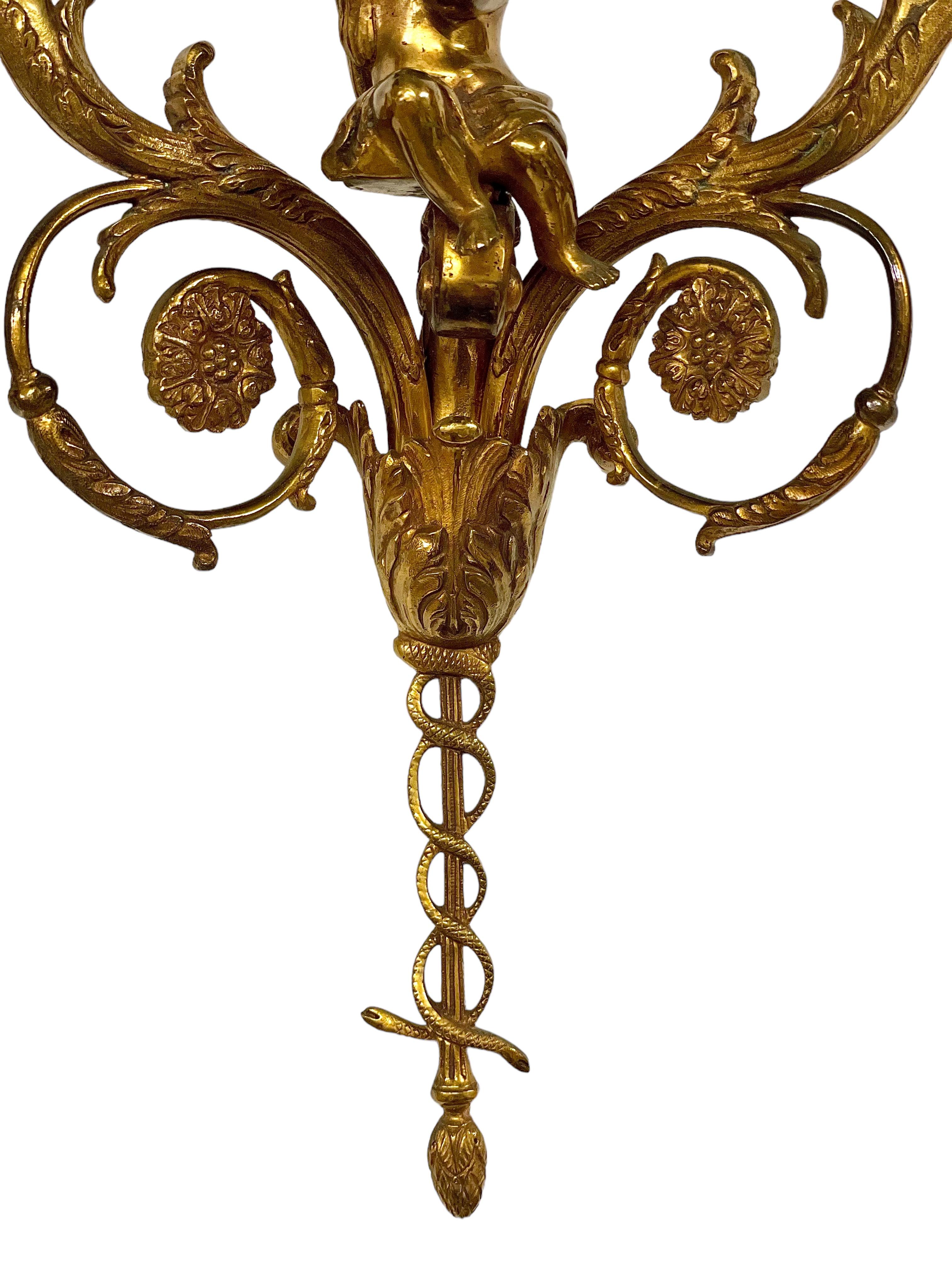Un par de apliques de pared de bronce dorado exquisitamente cincelados, cada uno con dos ramas de hojas que flanquean a un angelical querubín que toca la flauta. Las bandejas de goteo y las toberas ornamentadas rematan los dos brazos, que convergen