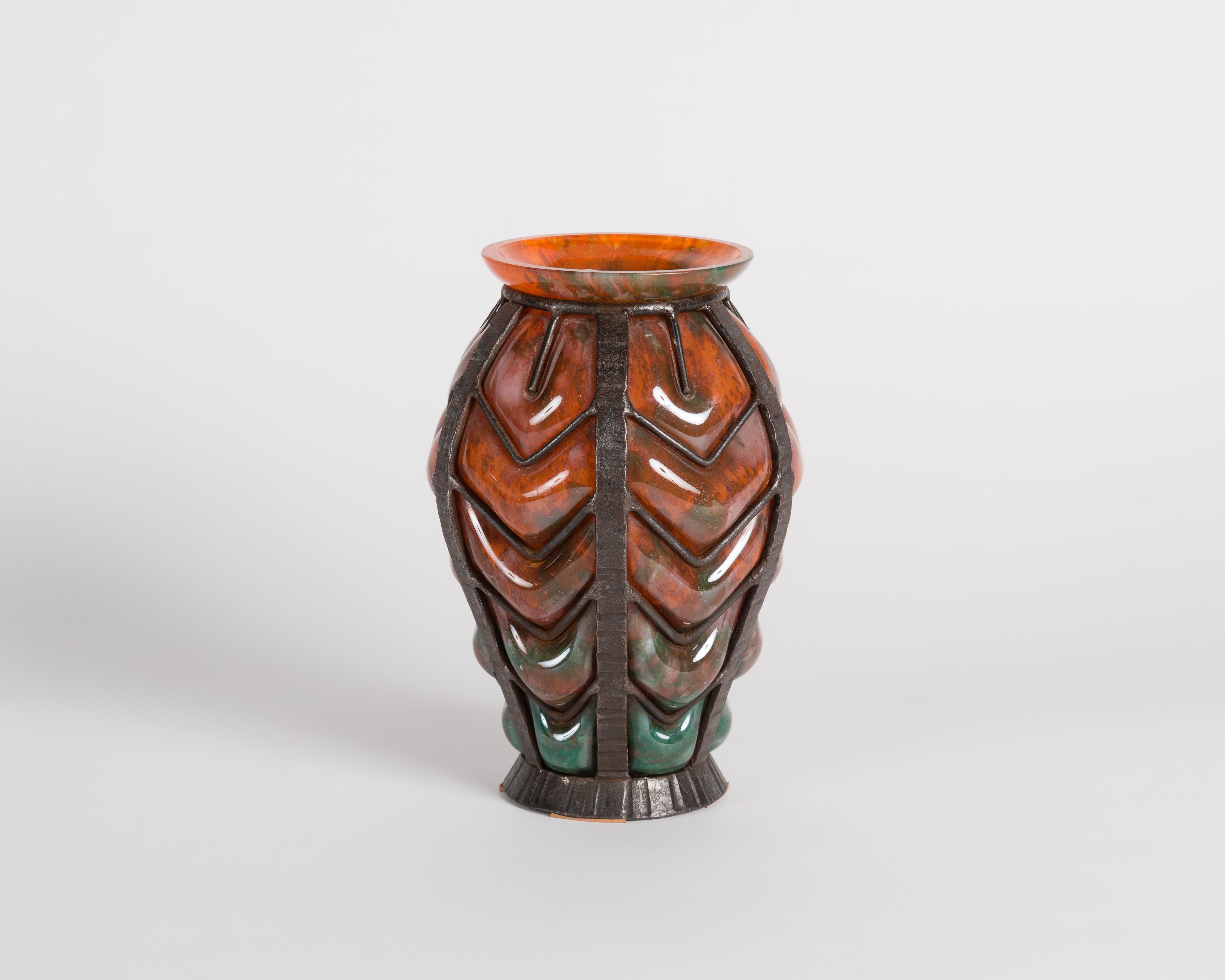 Ce vase du début de la période déco, attribué aux Verreries d'Art Lorrain, présente une couleur orange unique et est entouré d'un extérieur en fer forgé finement nervuré.
