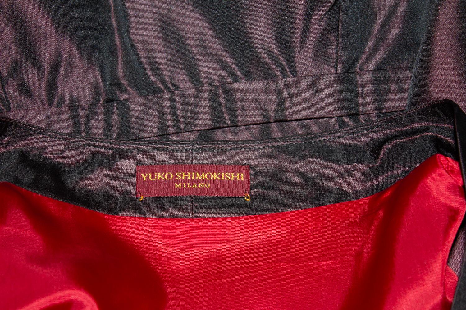 Un maravilloso vestido de cena/noche de Yuko Shimokishi de Milán. En un atractivo color berenjena con forro rojo, el vestido tiene escote en pico con  abertura lateral con cremallera . Tiene mangas hasta el codo y pliegues en la parte delantera.