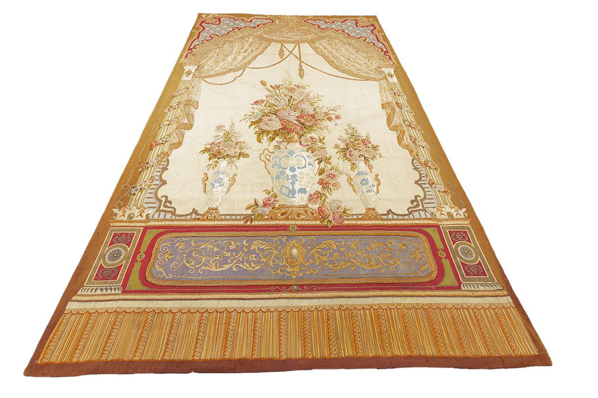 Cet exquis tapis à tissage plat d'Aubusson est un exemple frappant de l'art de la tapisserie française, présentant une somptueuse composition florale de nature morte dans un cadre théâtral et orné. Au cœur du dessin, une grande urne centrale