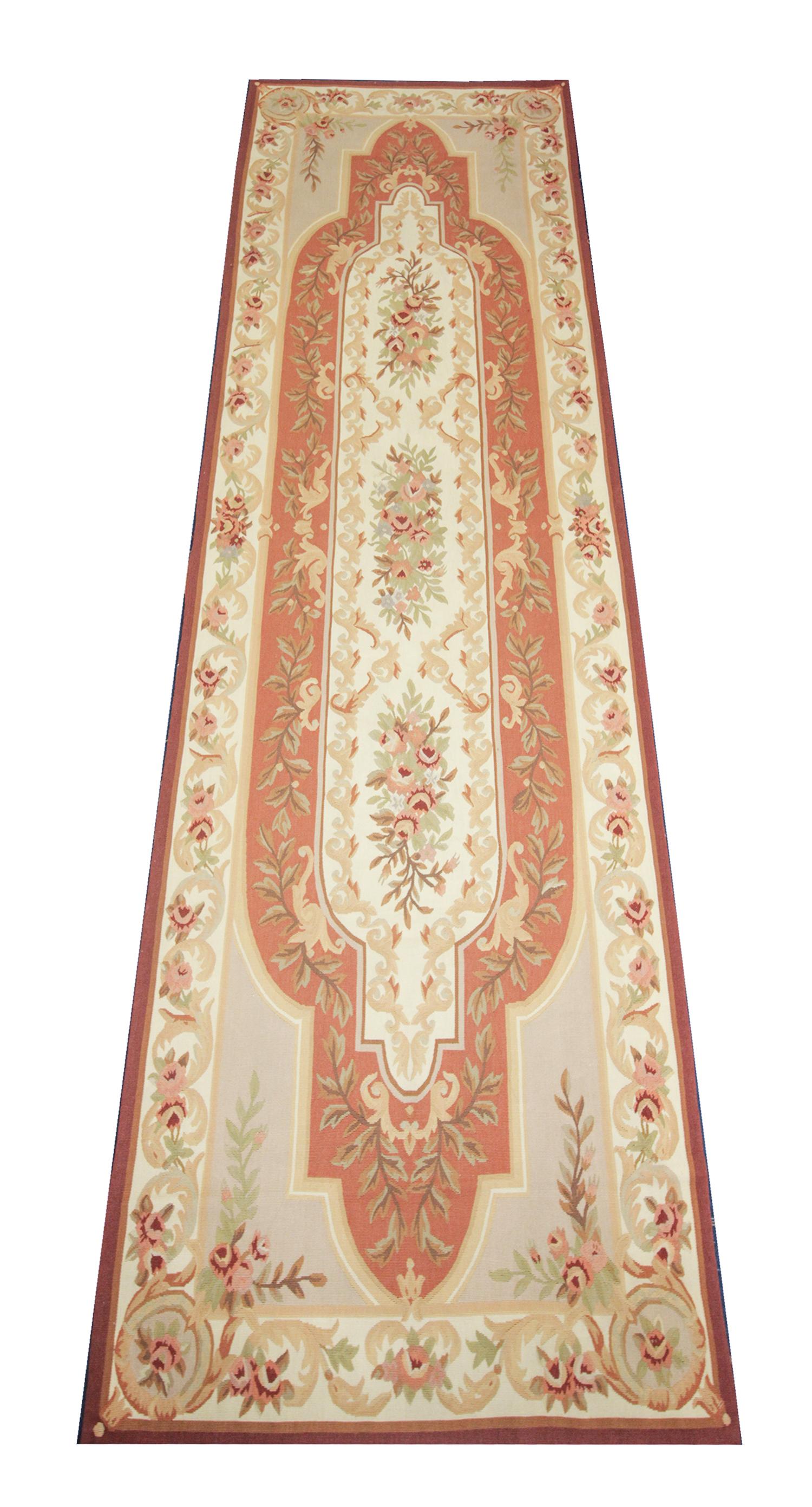 This handwoven oriental needlepoint rug was woven by hand in China in the early 21st century. The central design features a symmetrical floral pattern woven with orange, pink, beige, cream and green accents. This fine oriental rug runner combines