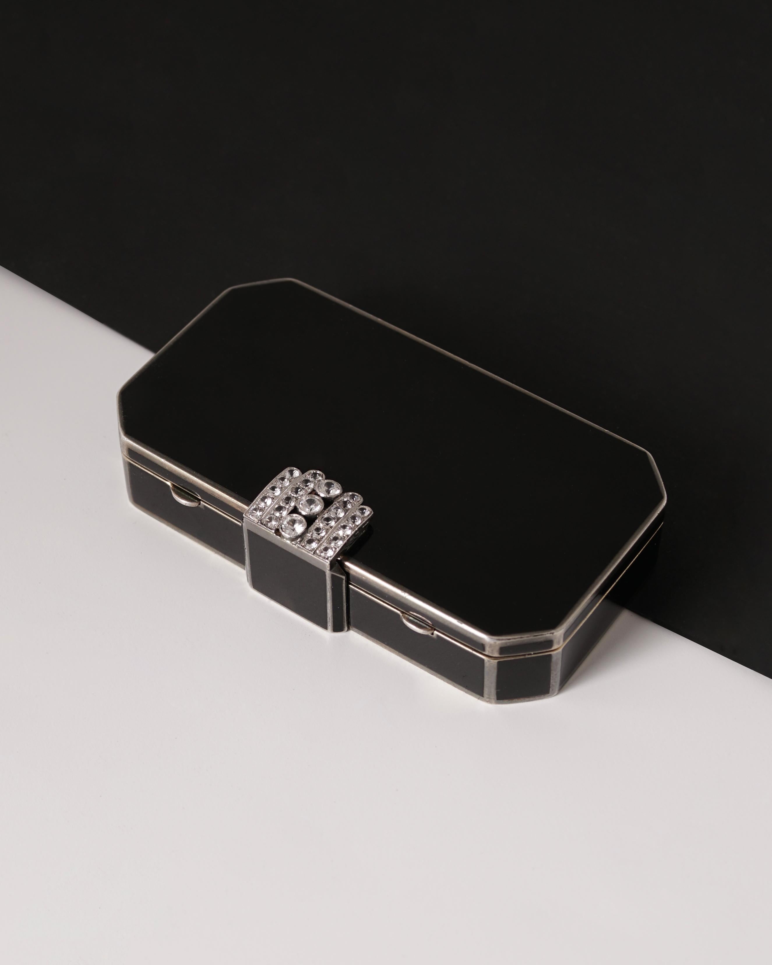 Auguste Peyroula
Compact:: Circa 1925
Or 18K:: argent:: émail noir:: fermoir coulissant serti de cinq bandes décoratives de diamants taillés en rose s'ouvrant sur un miroir dans le couvercle:: deux compartiments à charnière et un porte-rouge à