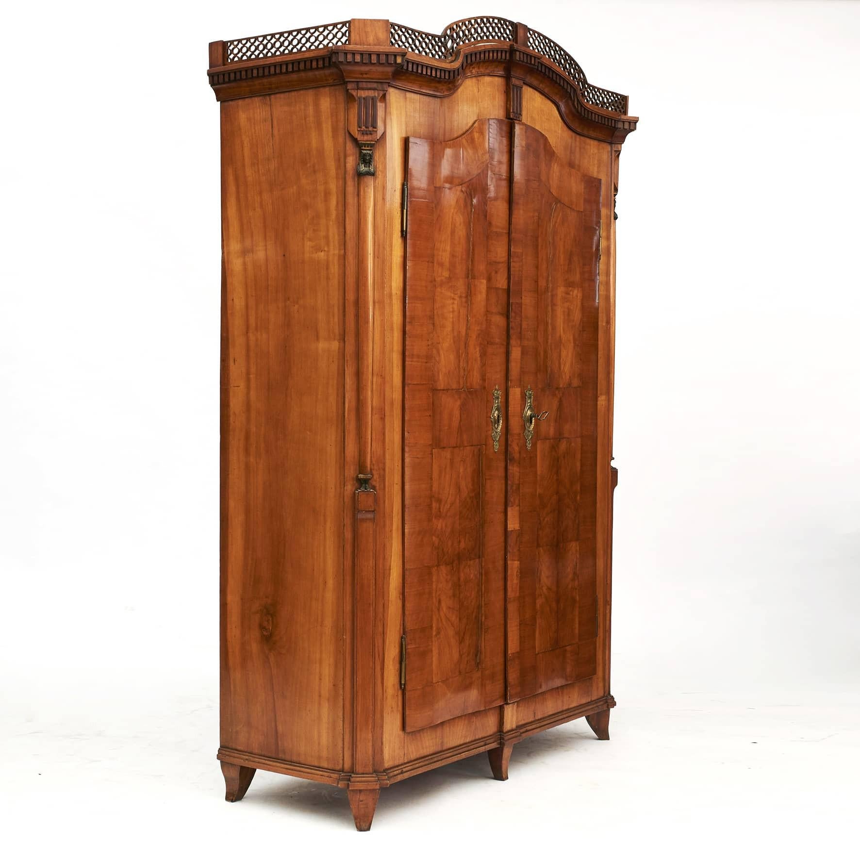 The two-door armoire or wardrobe has a cherry wood veneer with walnut fluting. Pierce-carved crown with dentil molding. Canted corners adorned with emerald green Egyptian caryatid pilasters with gold details. Doors with original fittings, key and