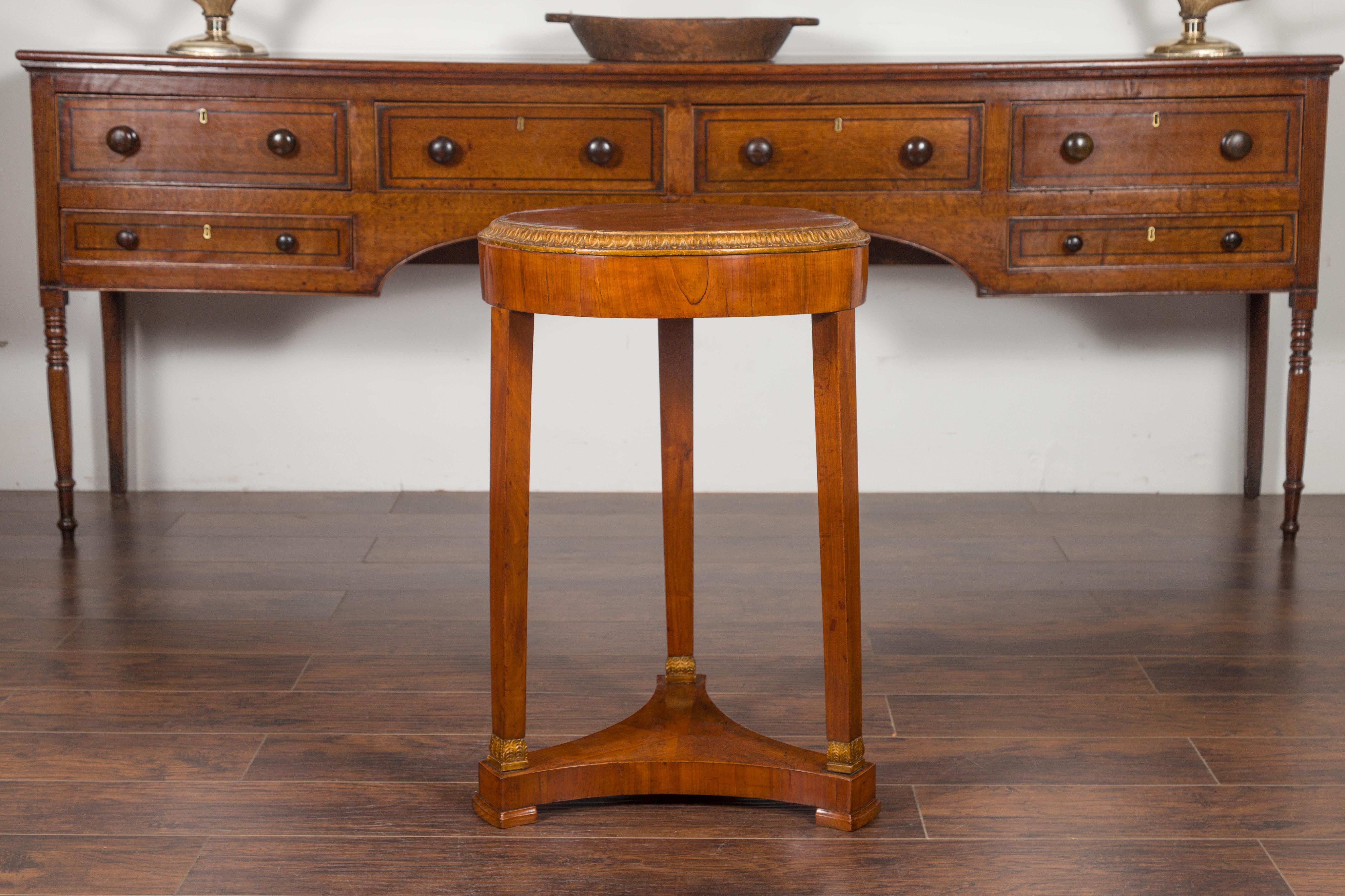 An Austrian Biedermeier period walnut tripod table from the mid-19th century, with gilt accents and tripartite shelf. Created in Austria during the second quarter of the 19th century, this Biedermeier table features a planked circular top surrounded
