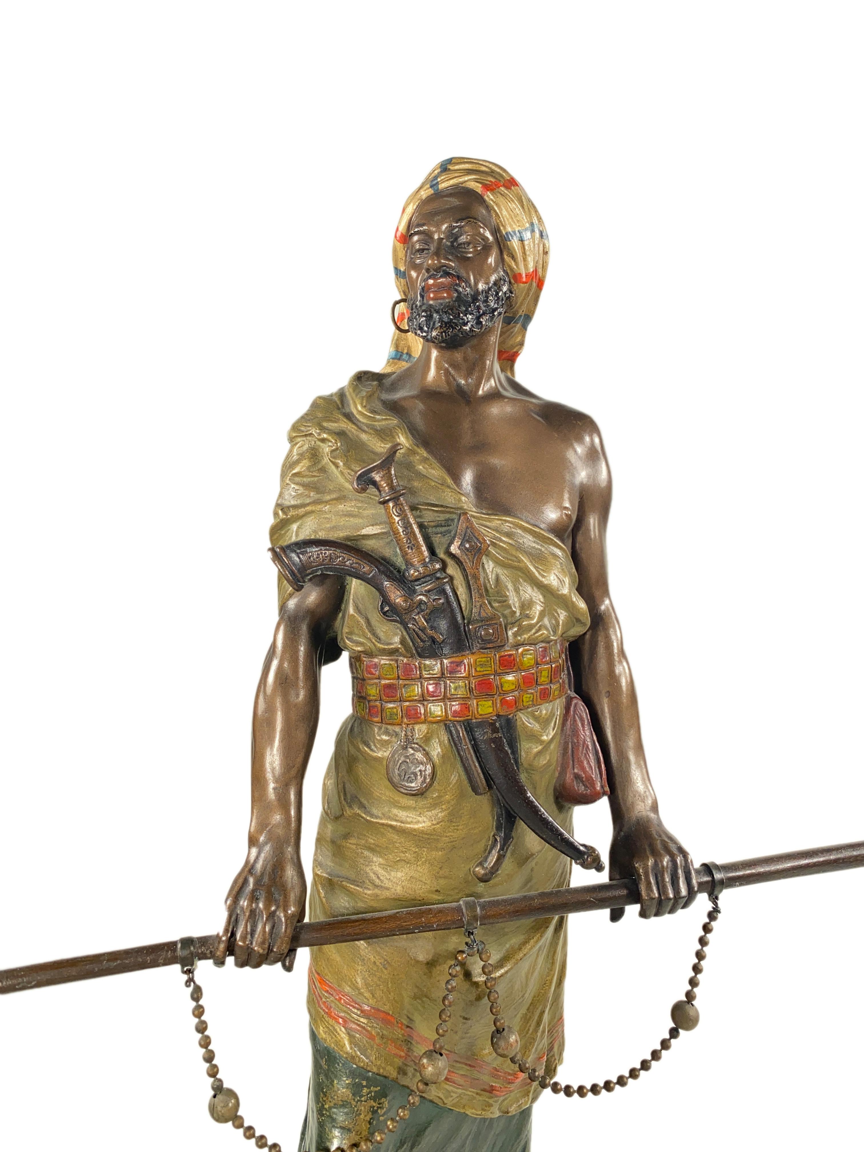 An Austrian Art Nouveau bronze Orientalist sculpture of An Arab Warrior attributed and most likely made by, Franz Bergman depicting a patinated, poly-chromed and cold- painted standing Arab warrior holding a chained rod in his hands, guns, and in