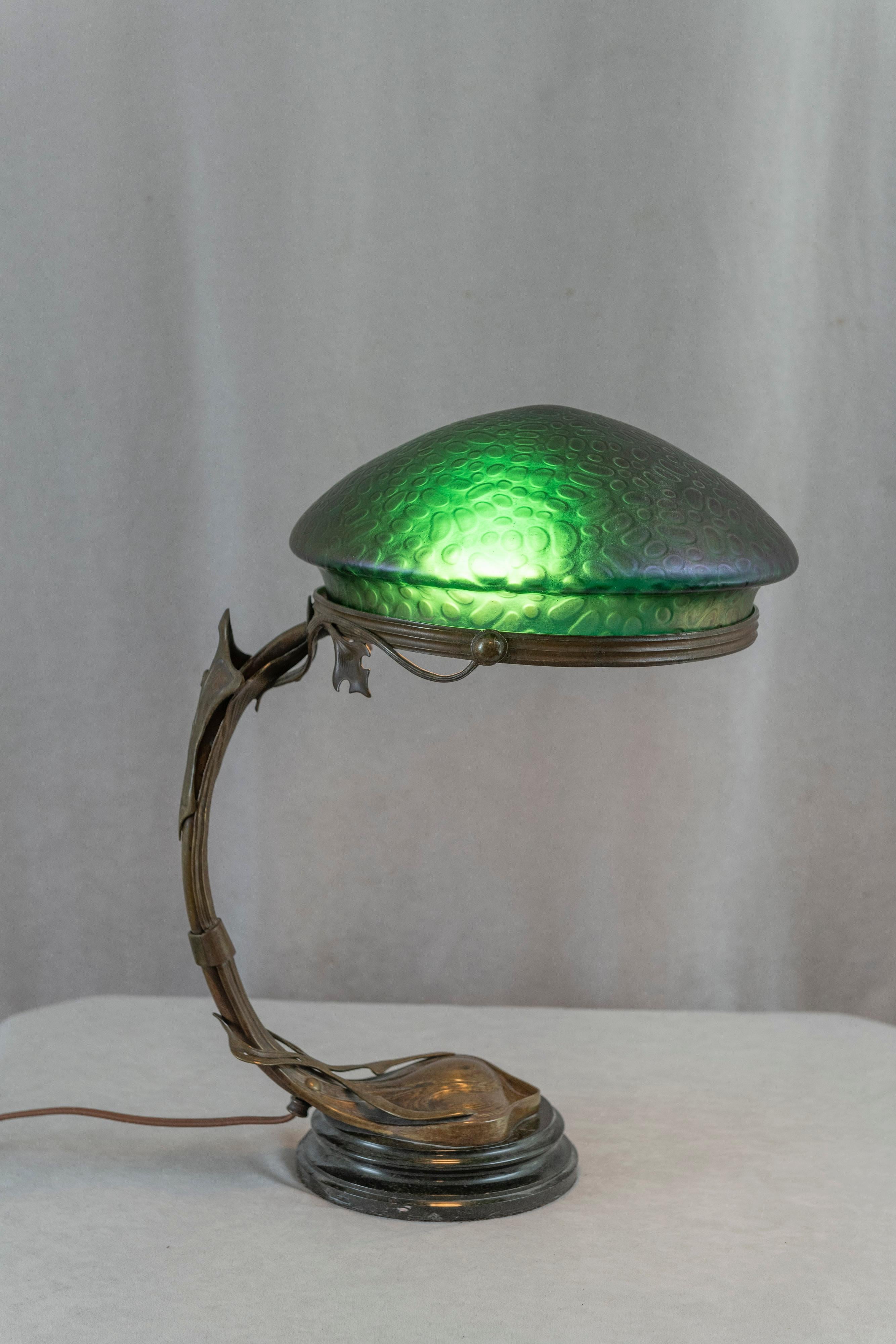 A fine example of Art Nouveau lighting. The bronze base is richly patinated and beautifully cast with an art nouveau design. The shade is a deep green when lit and purple and green when unlit. That's its iridescent quality. The shade is very much in