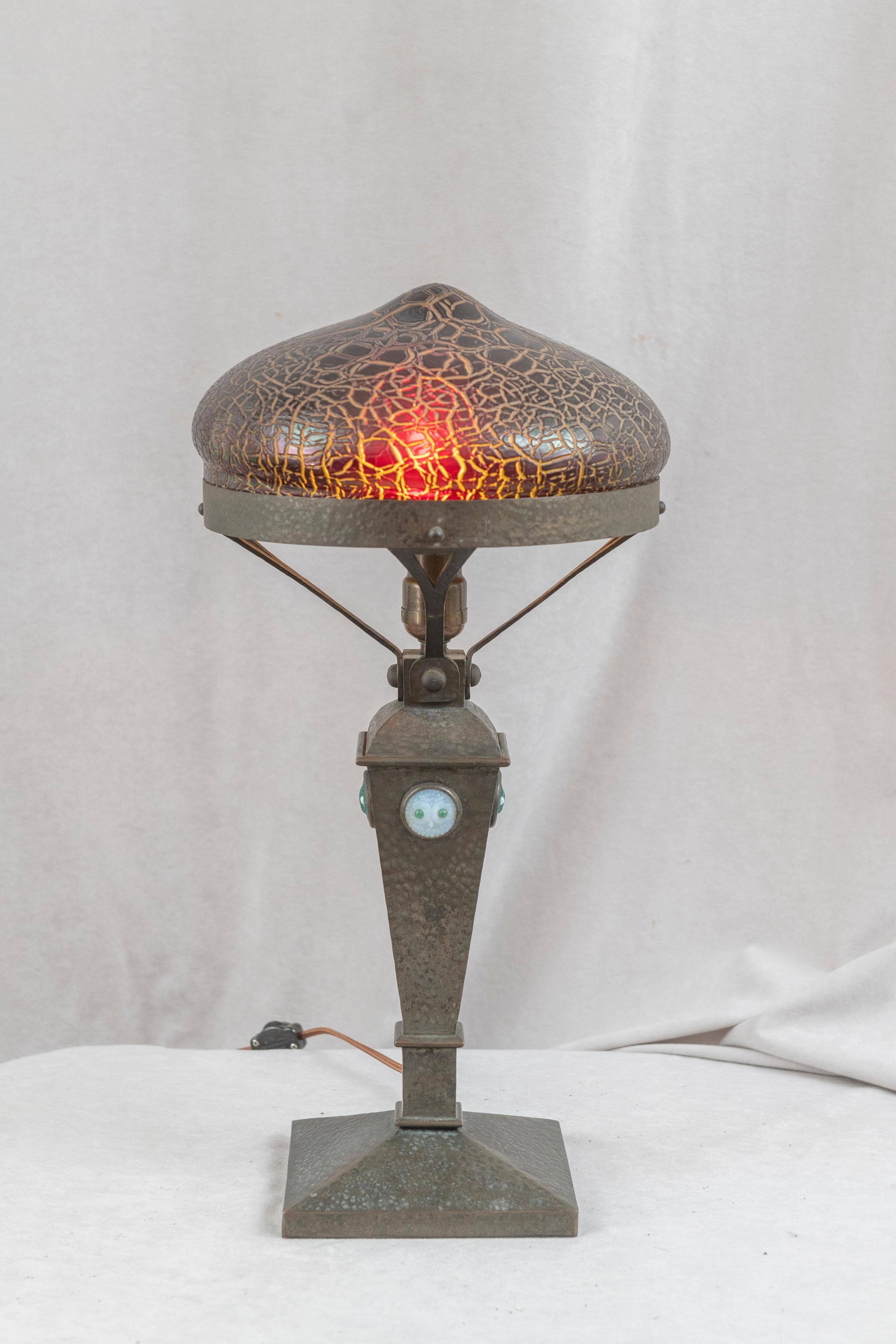This very special table lamp has many unique and distinct qualities. The richly patinated base, done in the arts & crafts tradition is hammered and on all 4 sides displays glass jewels with owls faces cast in the glass. 2 of the jewels are dark