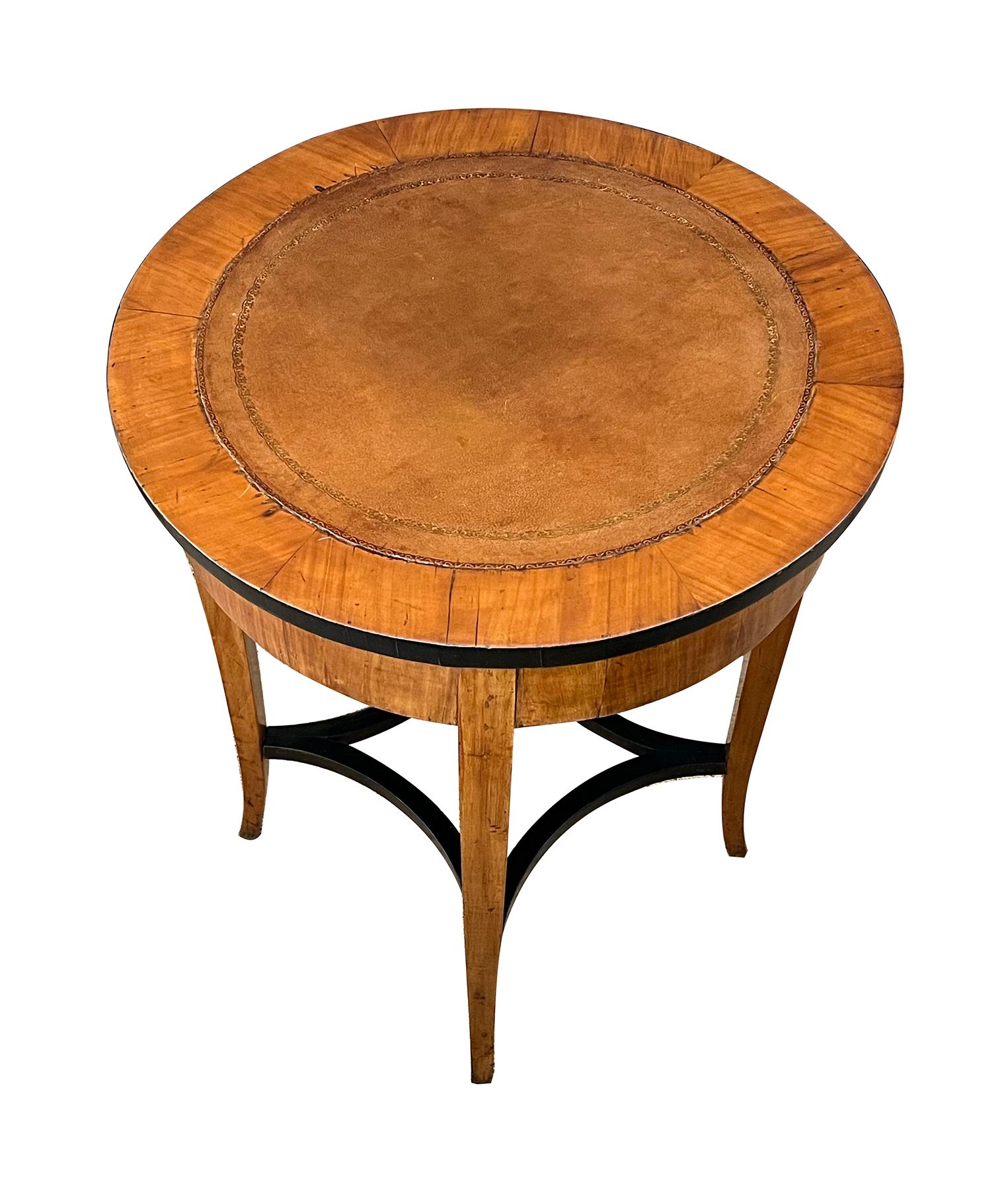 the round top with inset hand-tooled leather surface above a plain frieze apron; all raised on elegant splayed supports joined by an openwork ebonized stretcher; the top slides open to reveal fabric-covered compartments
