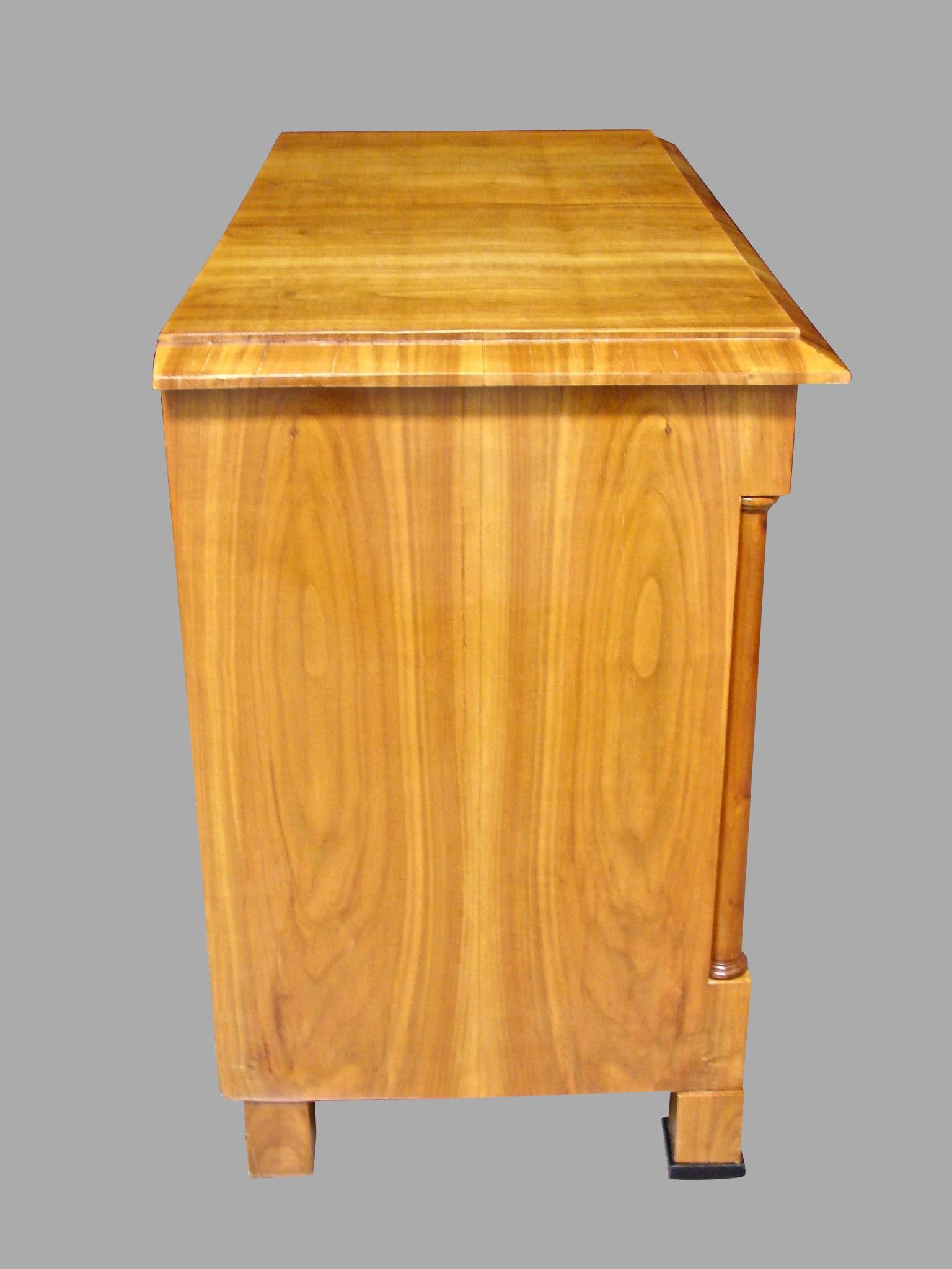 A good quality Austrian Biedermeier polished fruitwood (probably cherry) commode with figured well-matched veneers, the overhanging top above 3 long drawers framed by columns, each with an ebonized keyhole escutcheon, resting on block feet with