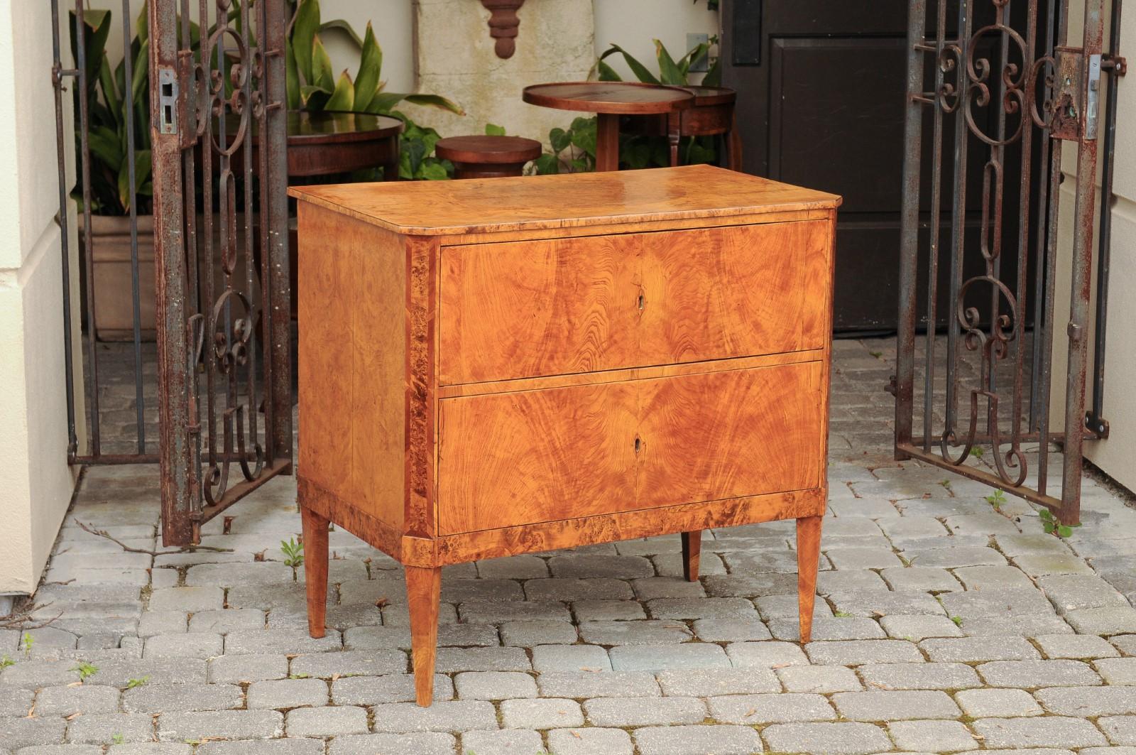 An Austrian Biedermeier period burled walnut veneered commode from the early 19th century, with bookmarked design, tapered legs and inlaid accents. Born in Imperial Austria during the first quarter of the 19th century, this exquisite Biedermeier