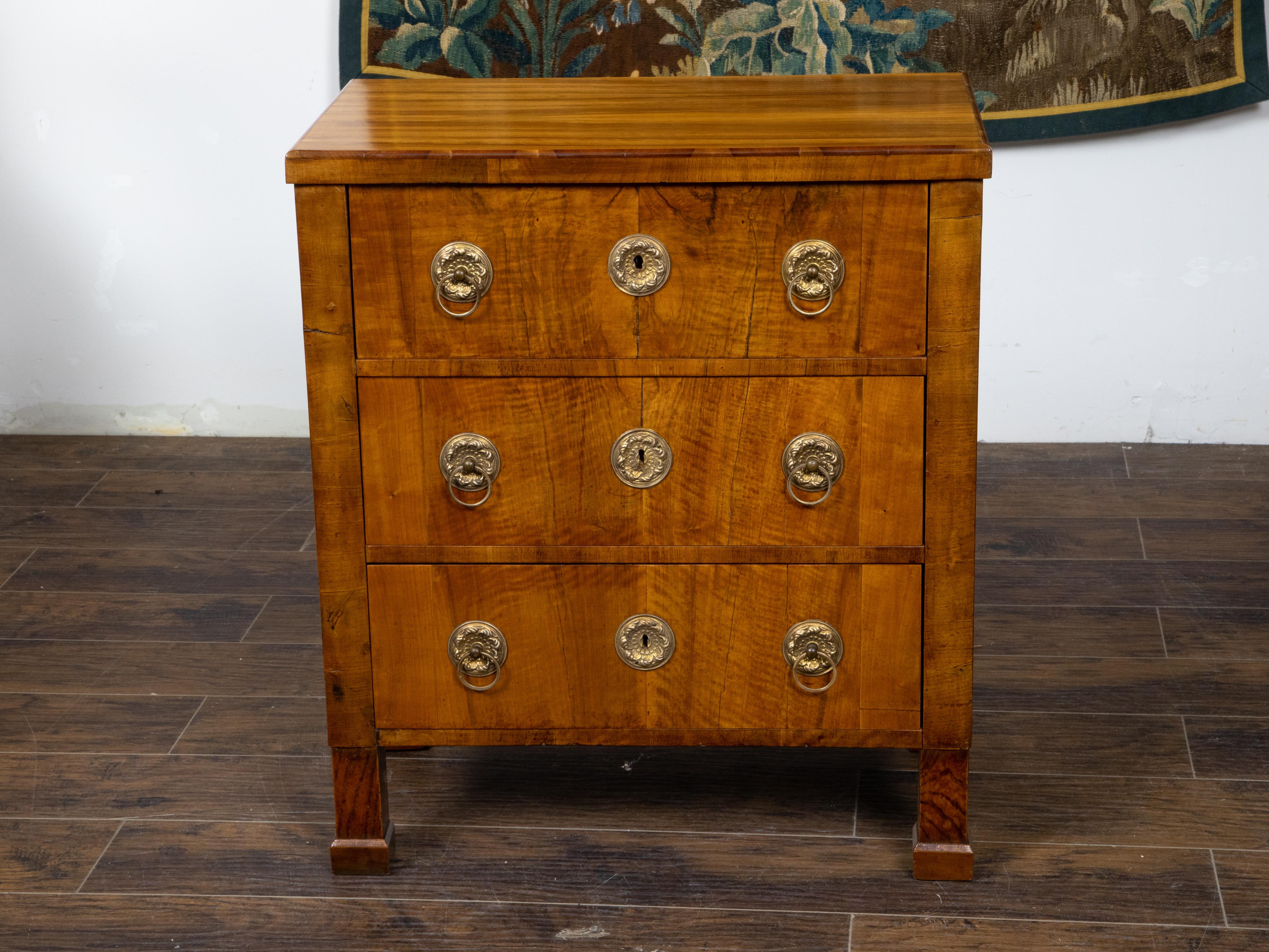 An Austrian Biedermeier period walnut veneered commode from the Mid-19th Century, with three drawers, butterfly veneer, ornate brass hardware and front block feet. Created in Austria during the second quarter of the 19th century, this walnut