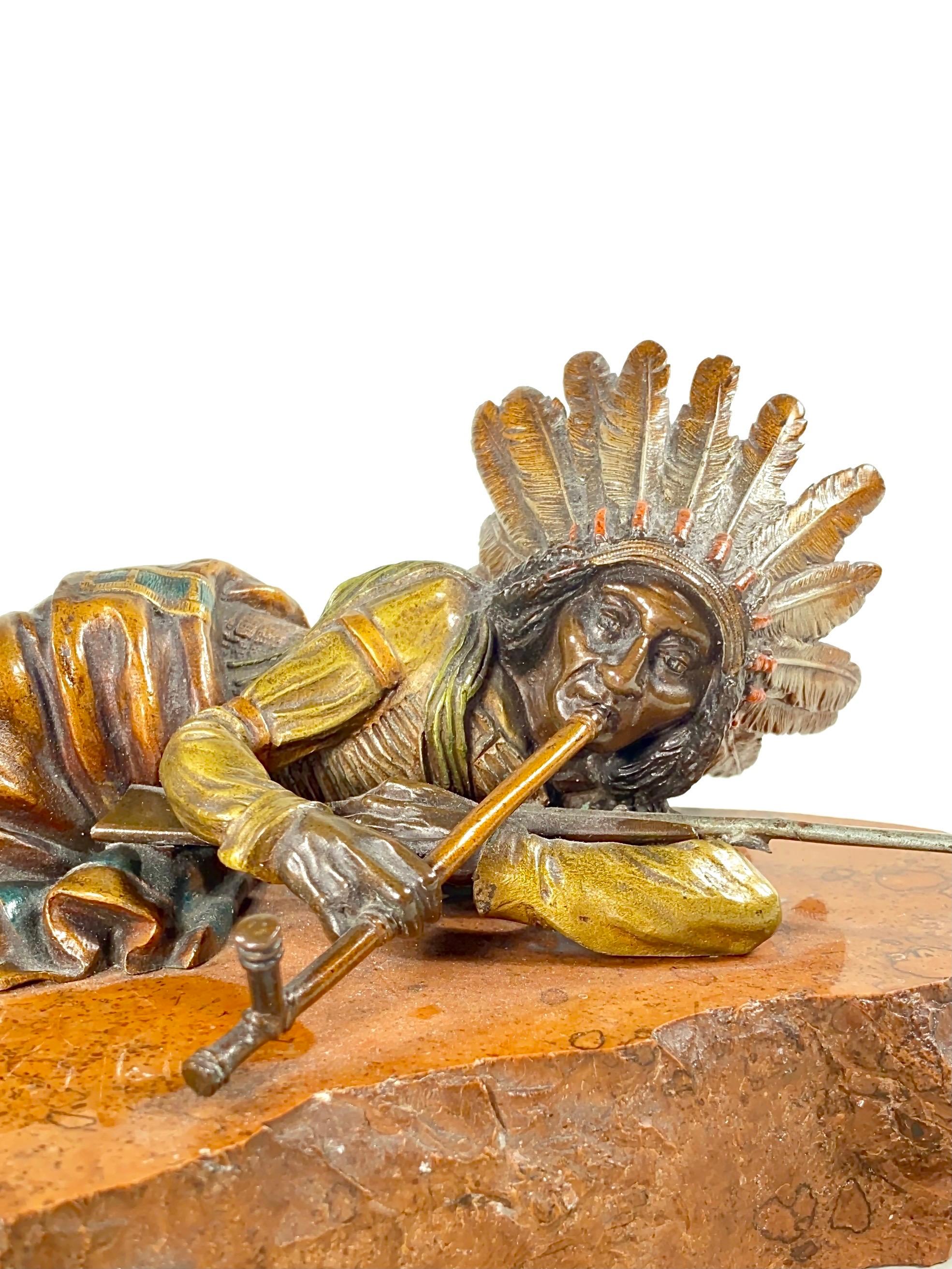 An Austrian Art Nouveau cold-painted cast bronze and stone sculpture of a Native American Indian Chief laying down with rifle cradled across and smoking a peace pipe a top its original petrified carved red stone base. The sculpture is impressed 