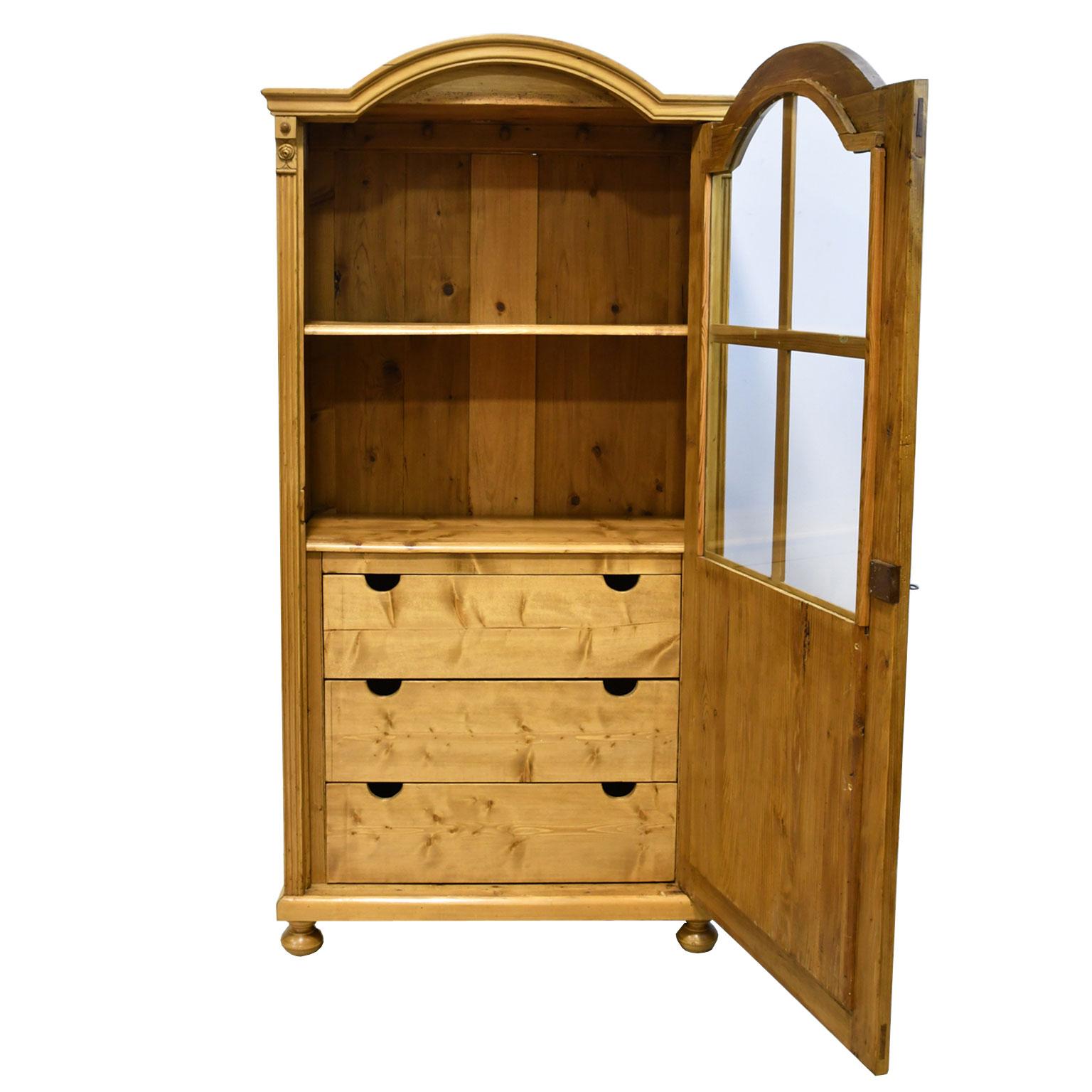 A lovely Austrian armoire or cabinet in pine with arched bonnet and single door with glazed upper panel opening to three large storage drawers and shelf. Cabinet rests on bun feet and has fluting on the front corners that are topped by a carved rose