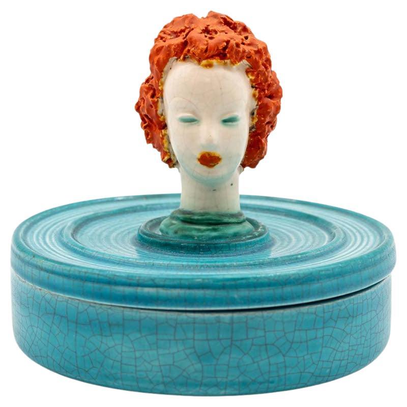 Un bol en céramique turquoise à glaçure craquelée et un couvercle assorti, tous deux finis dans une texture de glaçure craquelée. Le couvercle est surmonté d'une tête de femme sculptée qui sert de poignée. Les cheveux de la femme sont coiffés dans