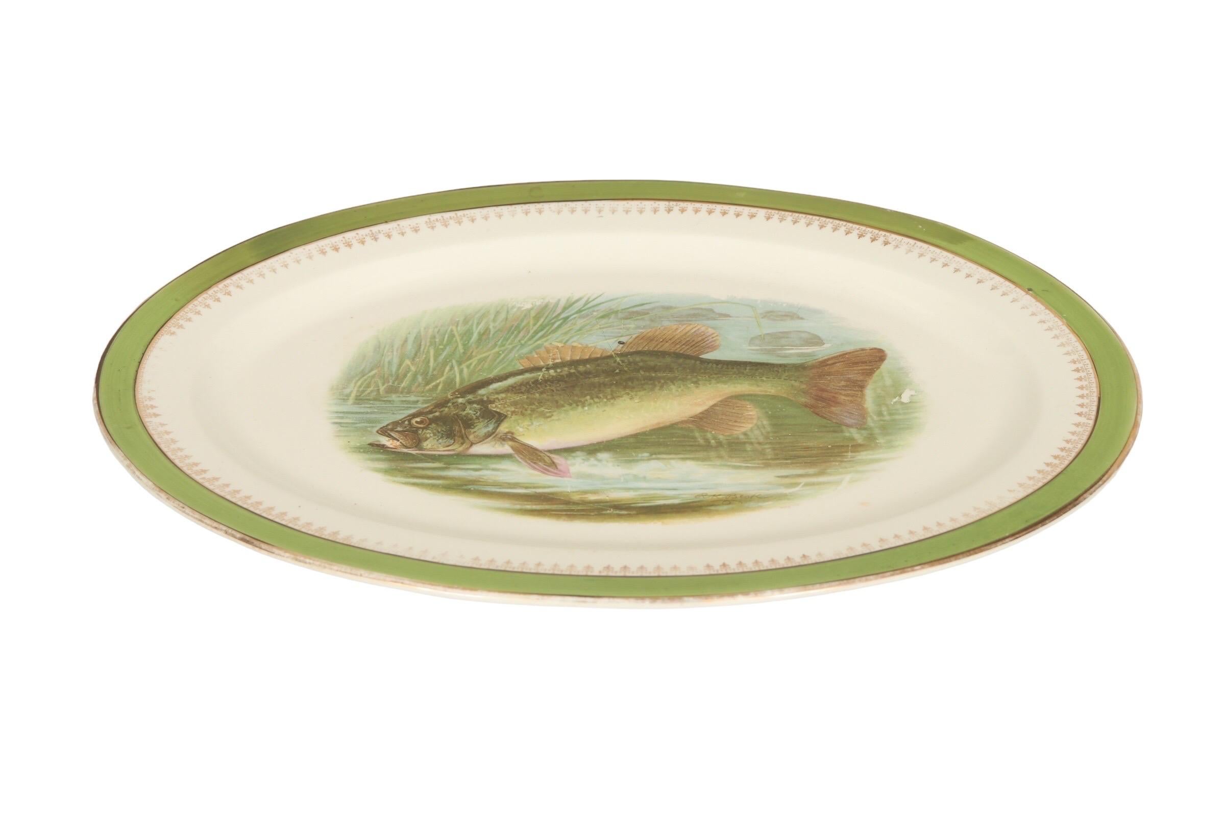 An Austrian ceramic serving platter decorated in the center with a transferware trout eating a shrimp. A gilt edge and lime green border are decorated with a gilt repeating motif. The artist's signature on the lower right reads “RK Boek”, marked