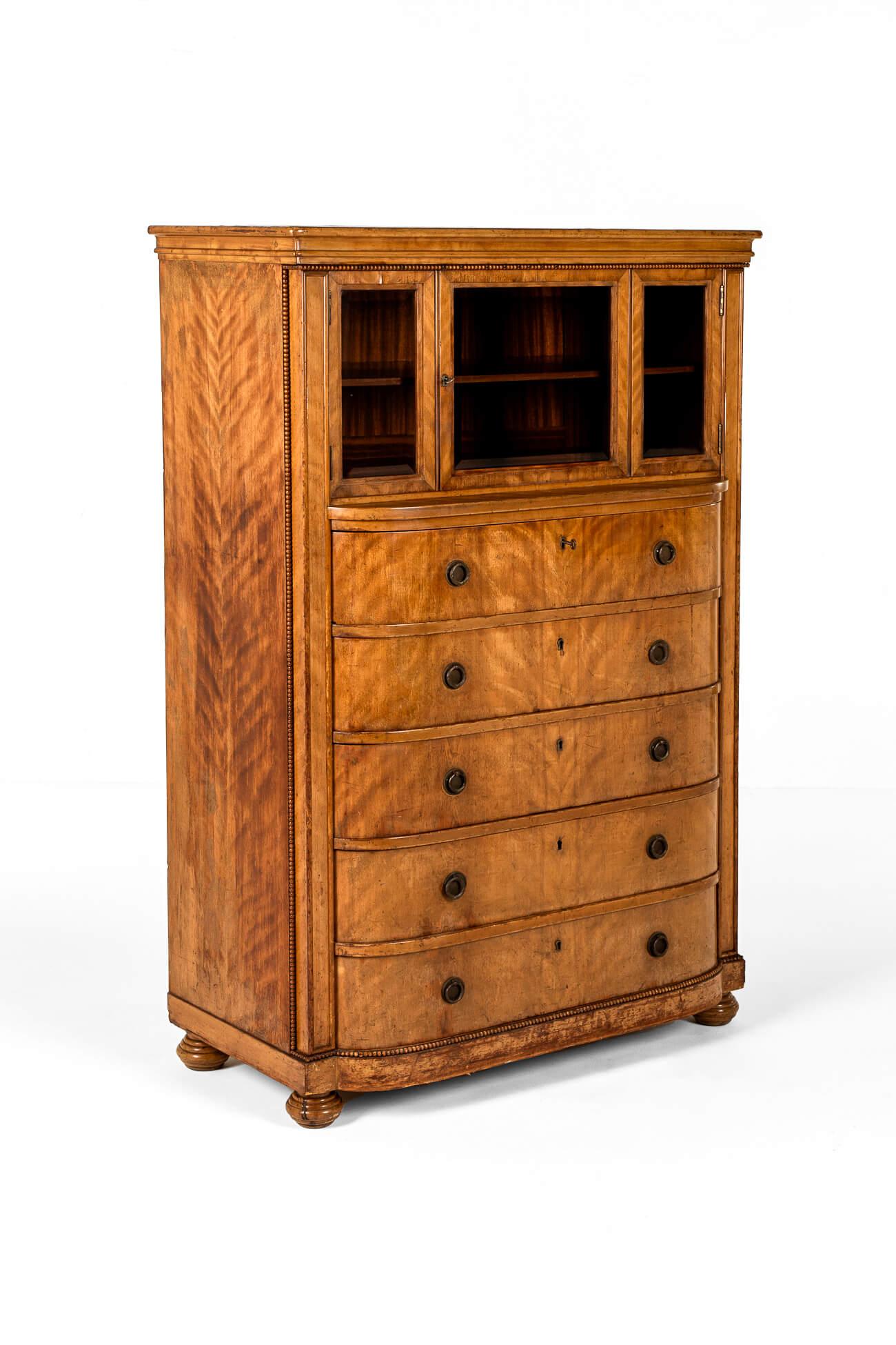 A tall glazed display cabinet over an arrangement of five long drawers in pine with satin wood veneers.

The molded stepped cornice features decorative beading with a three paneled glass display section to the top with original key and faux lock.