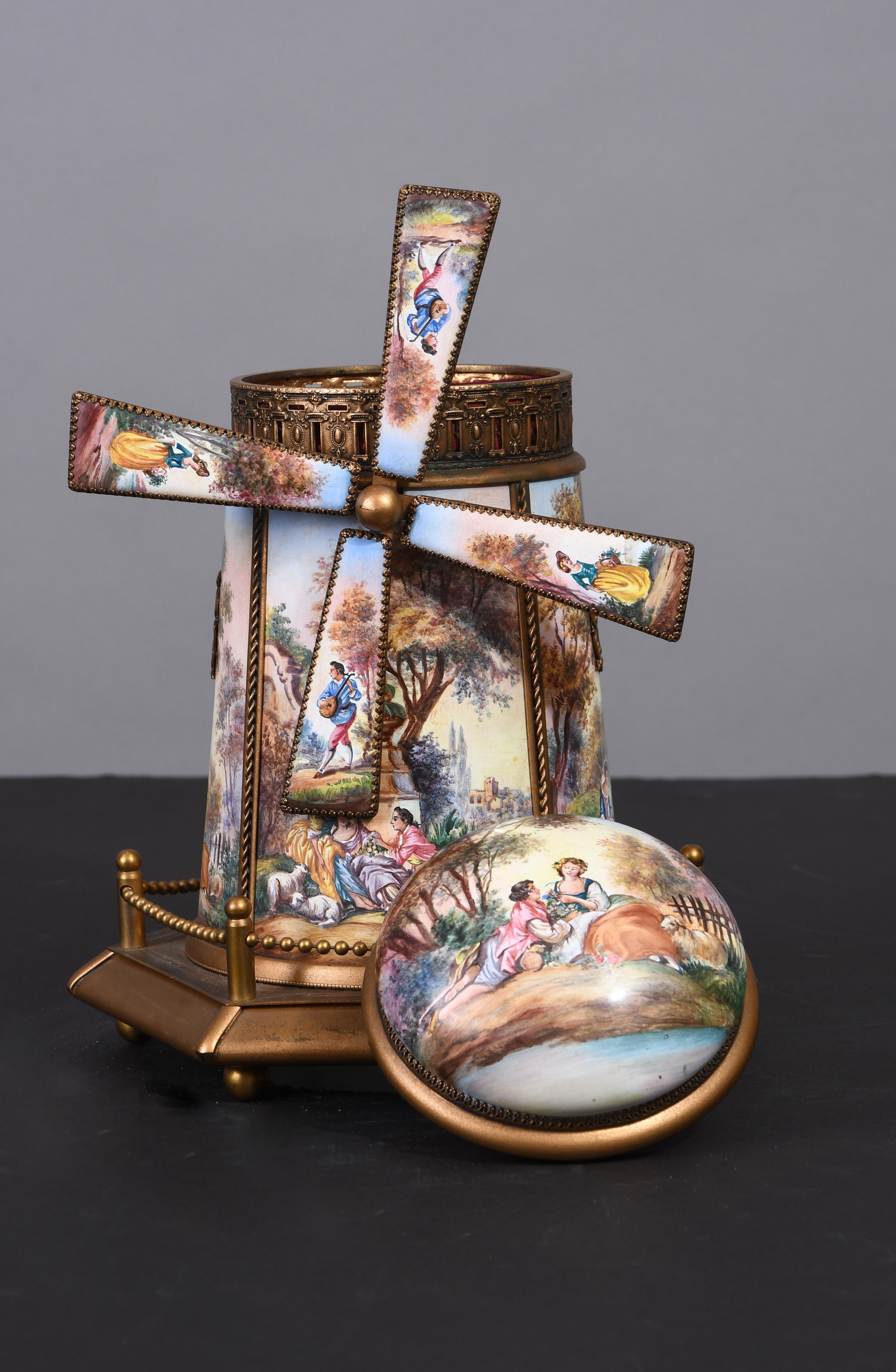 This windmill music box depicts different hand painted enamel panels of romantic courting couples in a medieval pastoral landscape. The windmill blades are adorned with shamrock prongs and show a gentleman playing the mandolin and a lady holding a