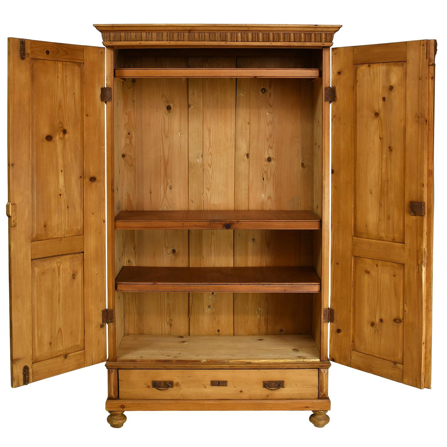 A lovely armoire in honey-colored pine with carved crown molding over two doors that open to three fixed shelves and rests on a base with one exterior drawer. Each door has two recessed panels, with carved acanthus leaf motif embellishing the top