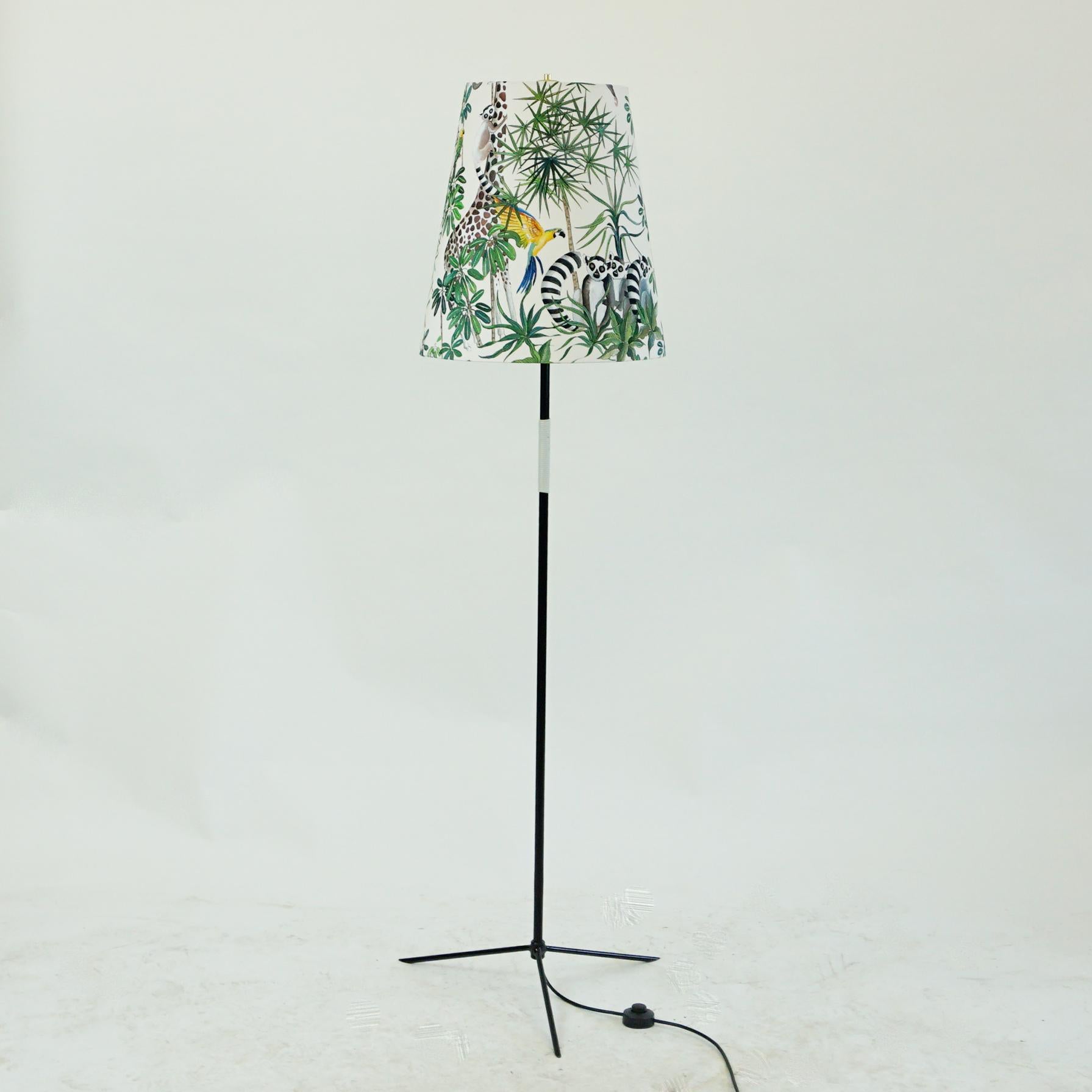 This charming Austrian midcentury floor lamp features a black lacquered tripod iron base and a brass final fitting on the top which holds the shade, which has been renewed and its fabric shows floral and animal Jungle decor. 
It has been designed